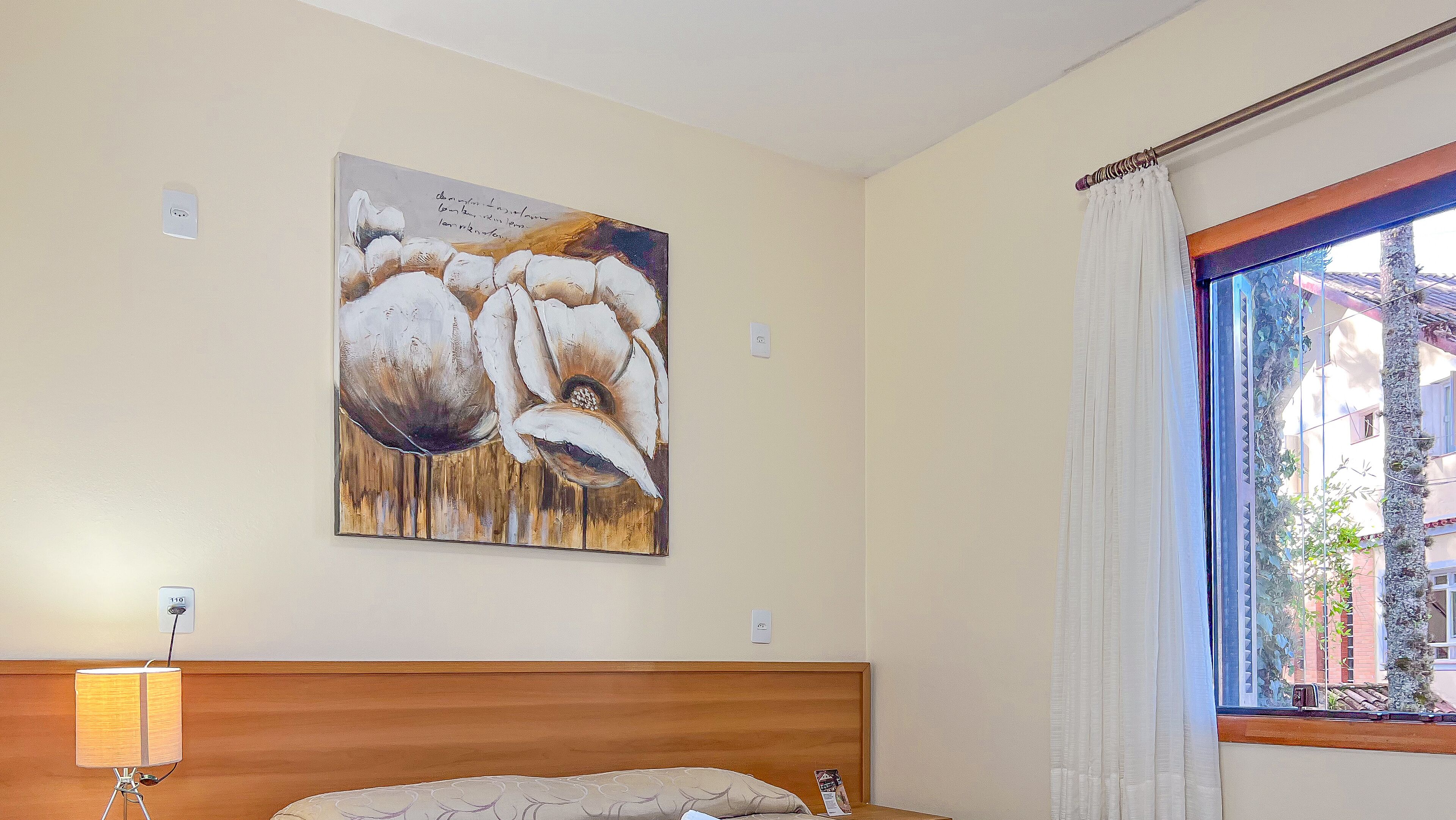 A bedroom with a wooden headboard, a painting of flowers on the wall, a bedside lamp, and a window with curtains.