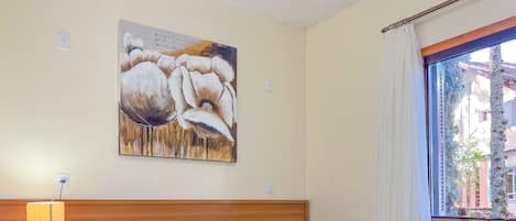 Un dormitorio con cabecera de madera, un cuadro de flores en la pared, una lámpara de noche y una ventana con cortinas.
