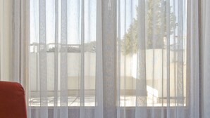 A room with a large window covered by sheer curtains, a red sofa with a yellow pillow, and a view of a building and trees outside.