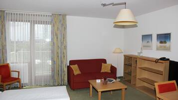 A hotel room with a bed, a red sofa, a wooden table, a desk, a lamp, and a window with curtains.