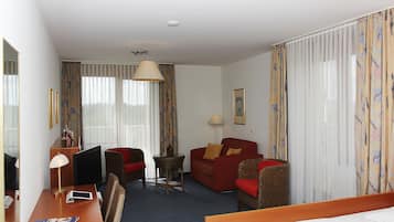 Camera d'albergo con un letto, una scrivania con un computer, un divano rosso, una poltrona in vimini, un tavolino con una lampada e un'ampia finestra con tende.