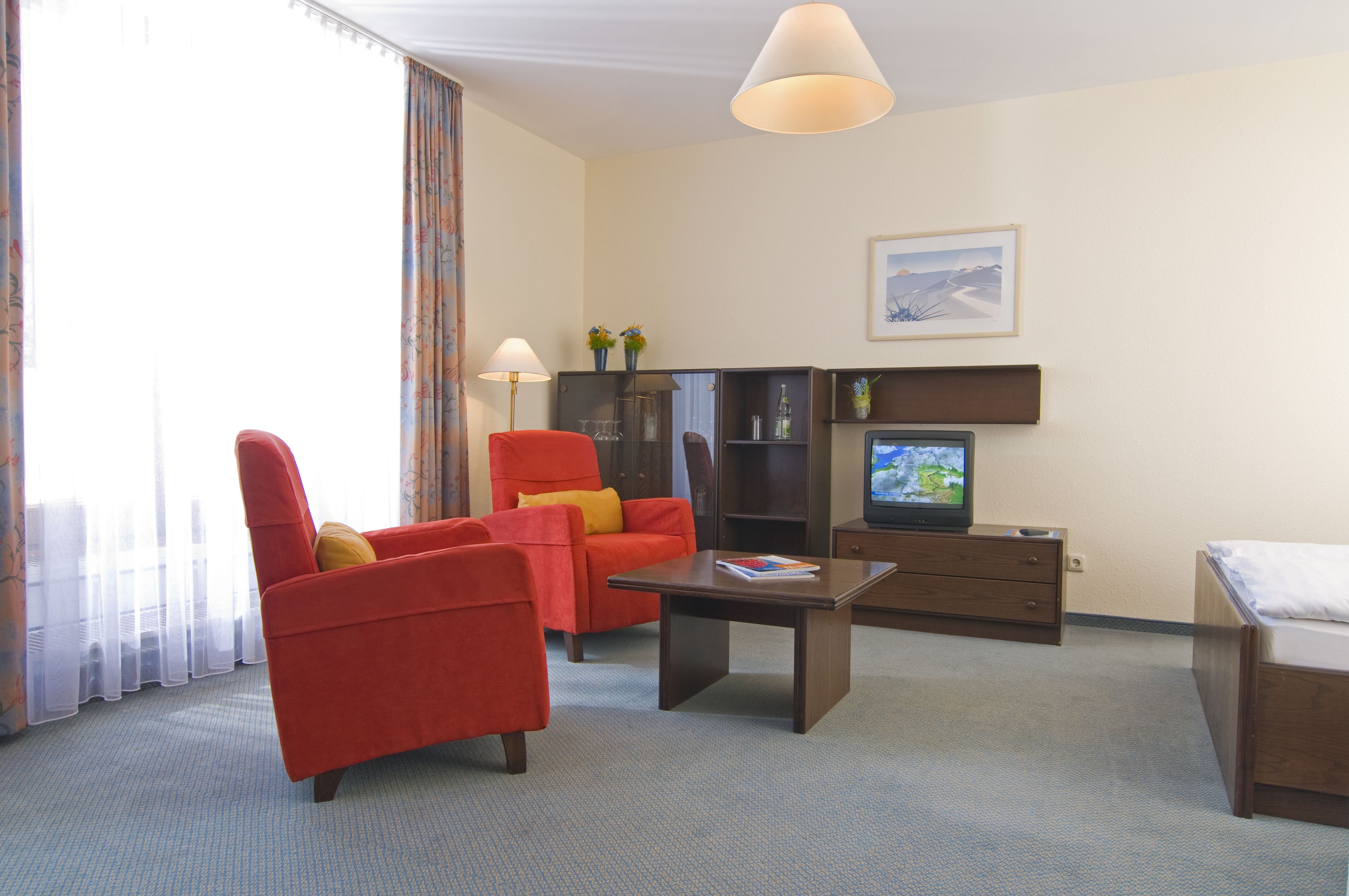 Una habitación de hotel con un sillón rojo, una mesa de centro de madera, un televisor, una cama y un cuadro en la pared.