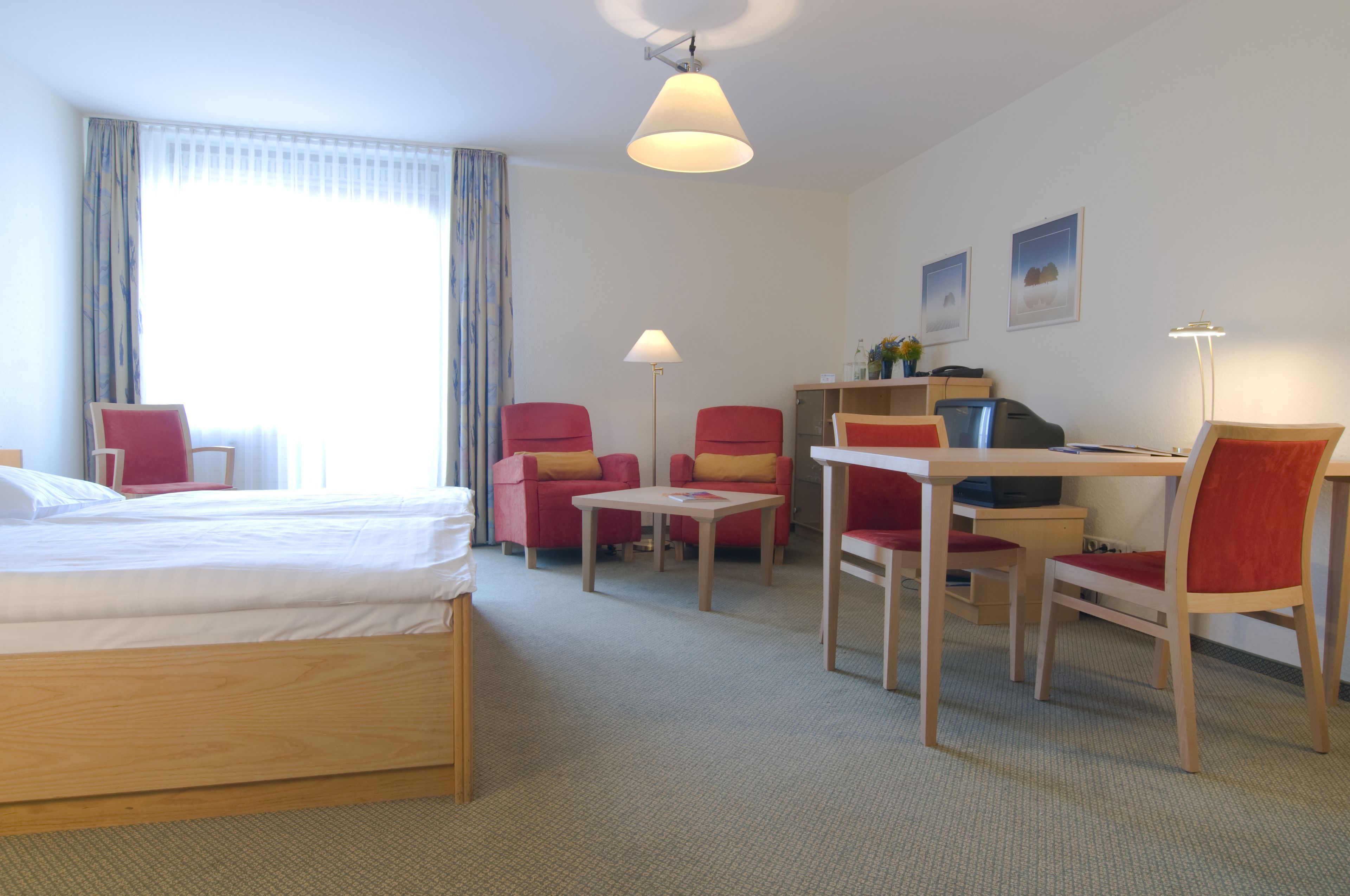 A hotel room with a bed, a desk with a chair, a small table, and two red chairs.