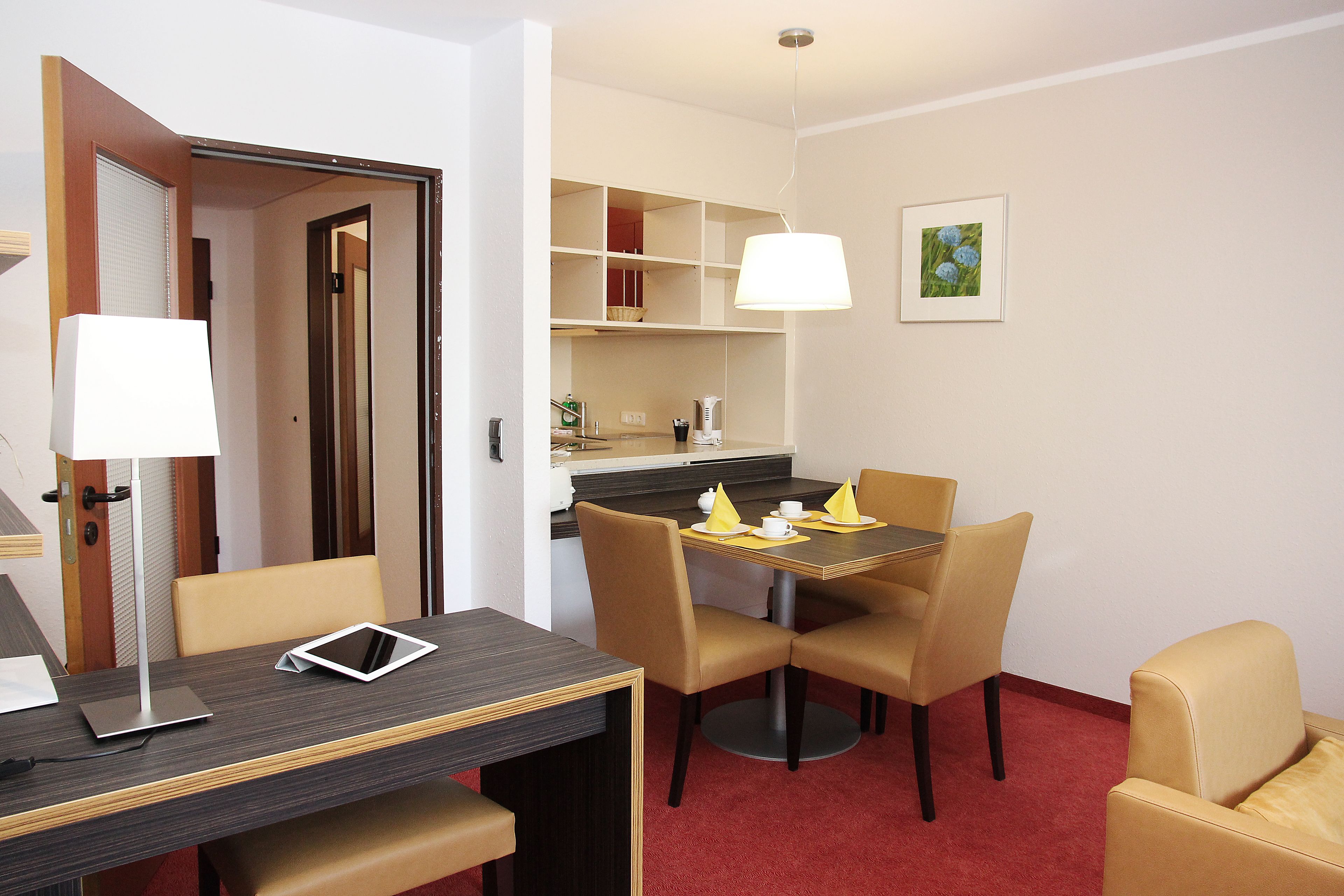 Et hotelværelse med spisebord, stole, en bærbar computer, en tablet, en lampe, et rødt tæppe, et tekøkken og et billede i ramme på væggen.