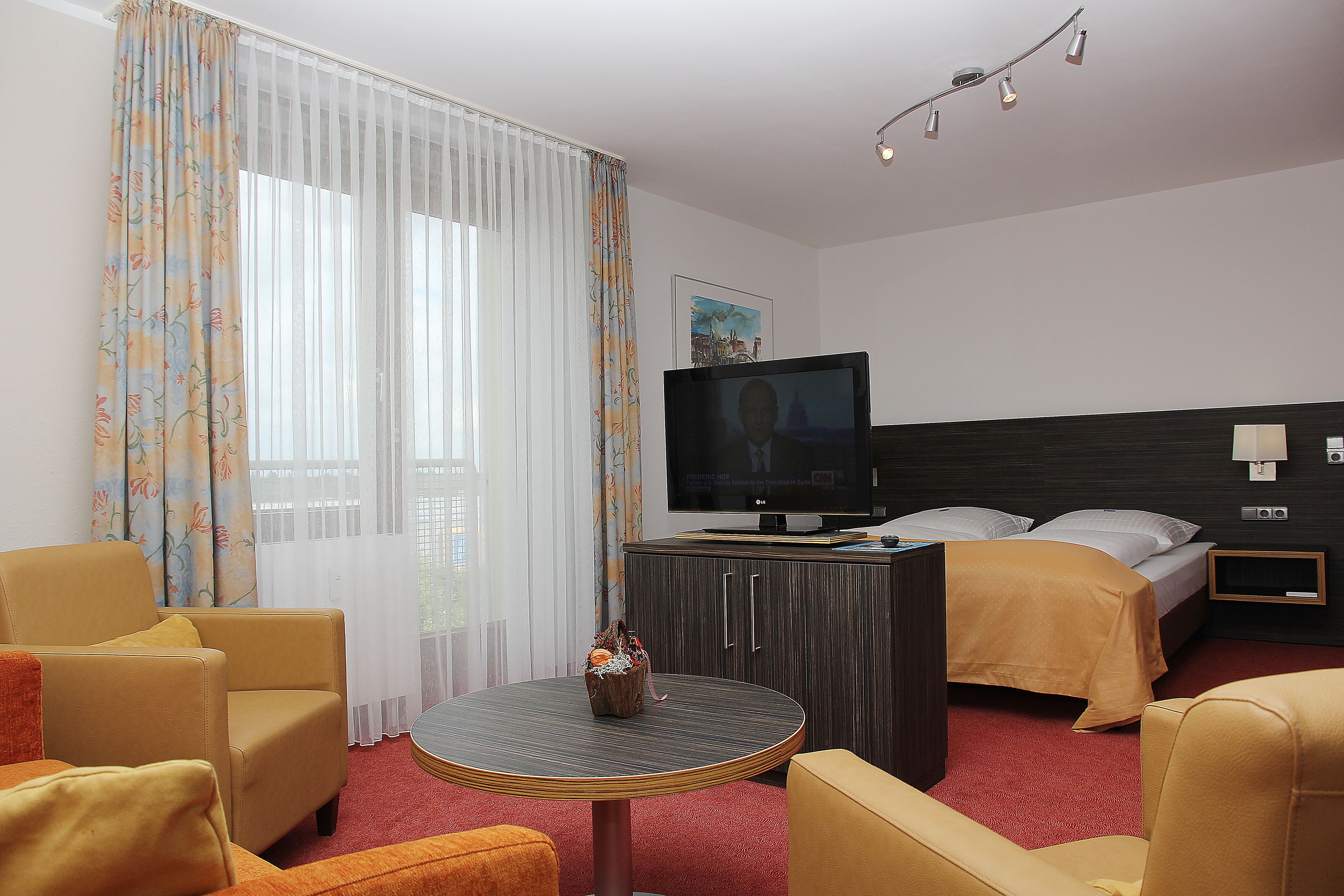 Una habitación de hotel con una cama, televisión, una mesa redonda con un florero y dos sillones.