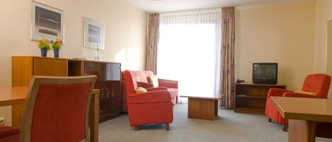 Habitación de hotel con zona de comedor, sofá, televisor y ventana con cortinas.
