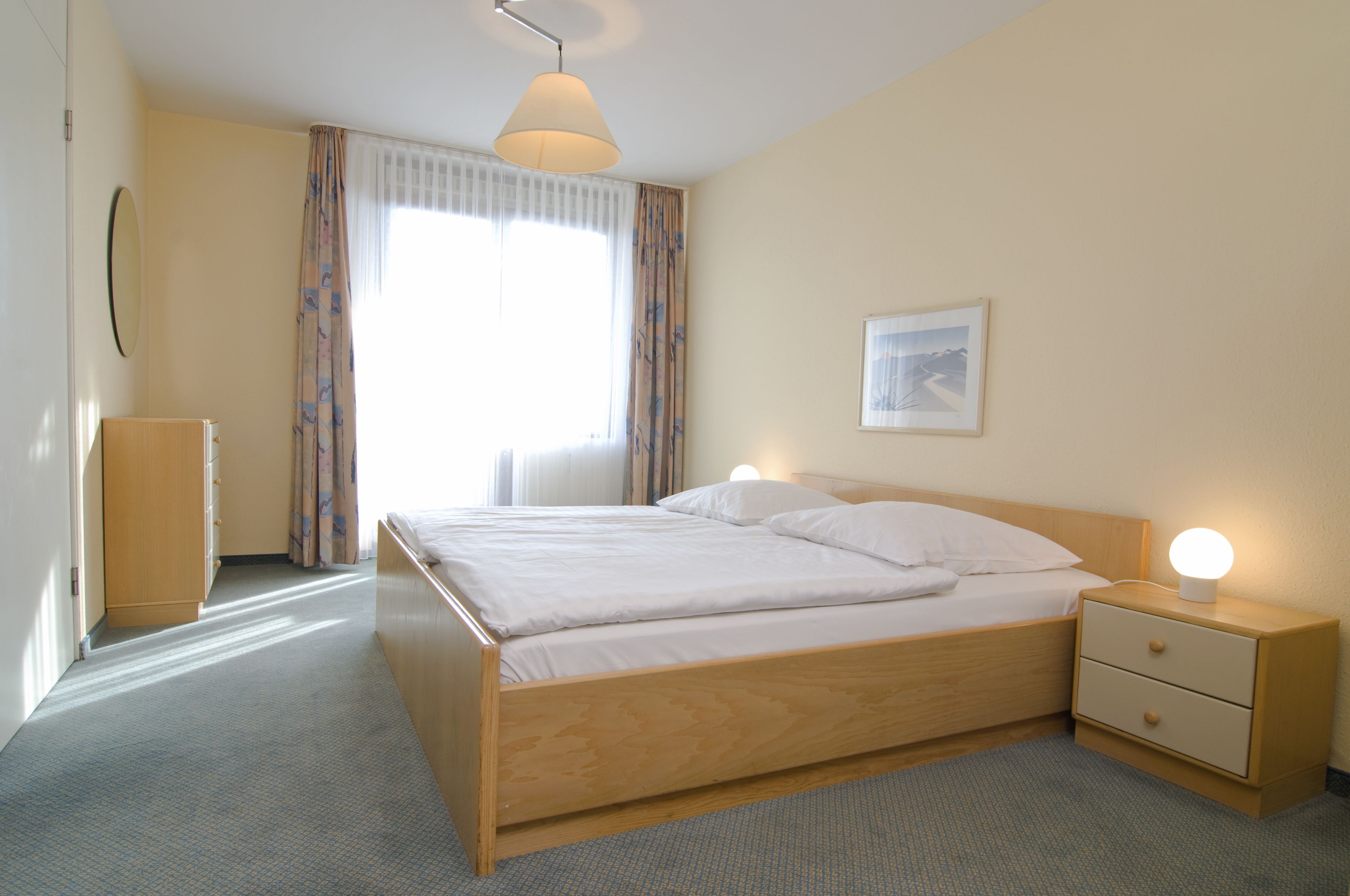 Une chambre d’hôtel bien rangée, avec un lit en bois, une table de chevet avec une lampe, une armoire et une fenêtre avec des rideaux.