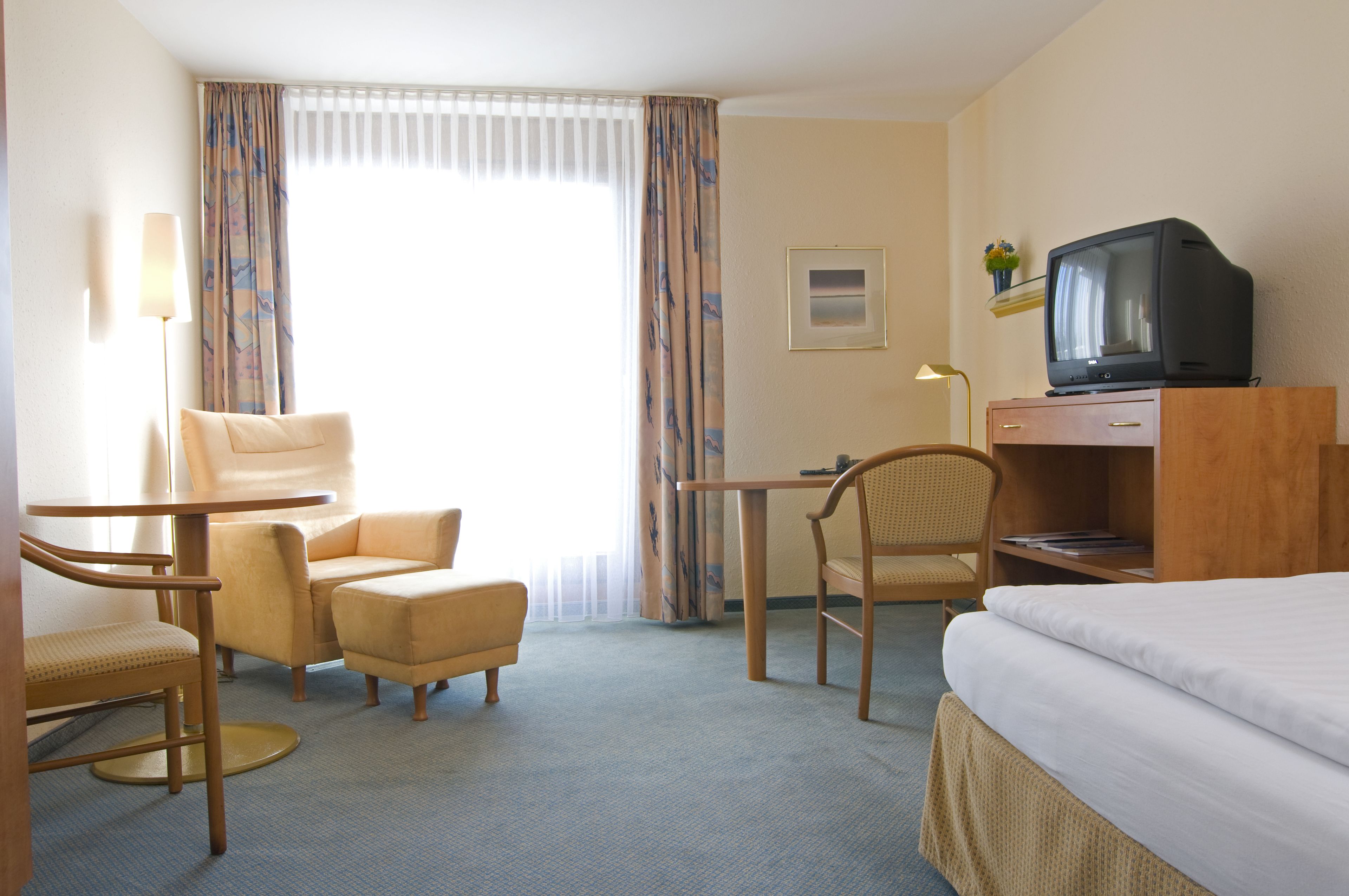 Una habitación de hotel con una cama, dos sillones, una mesita, un televisor sobre un mueble de madera y una ventana con cortinas.