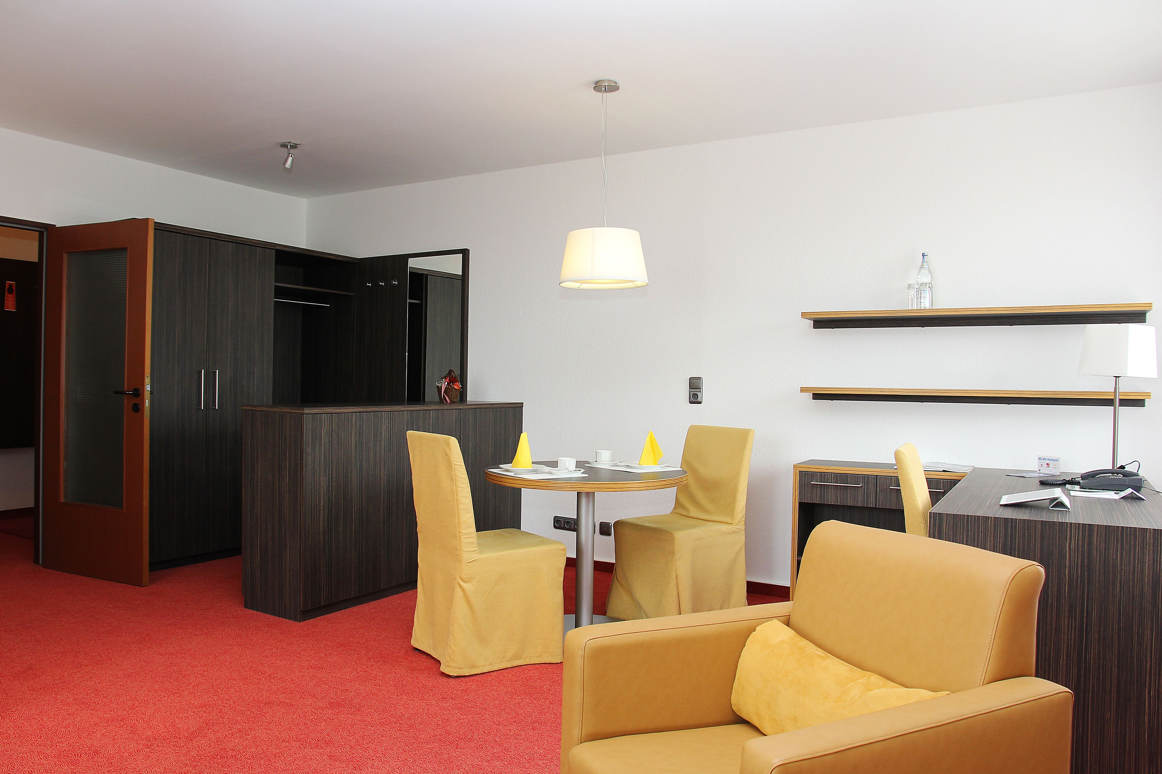 A hotel room with a red carpet, a round dining table with yellow chairs, a sofa, a desk with a chair, and a hanging lamp.