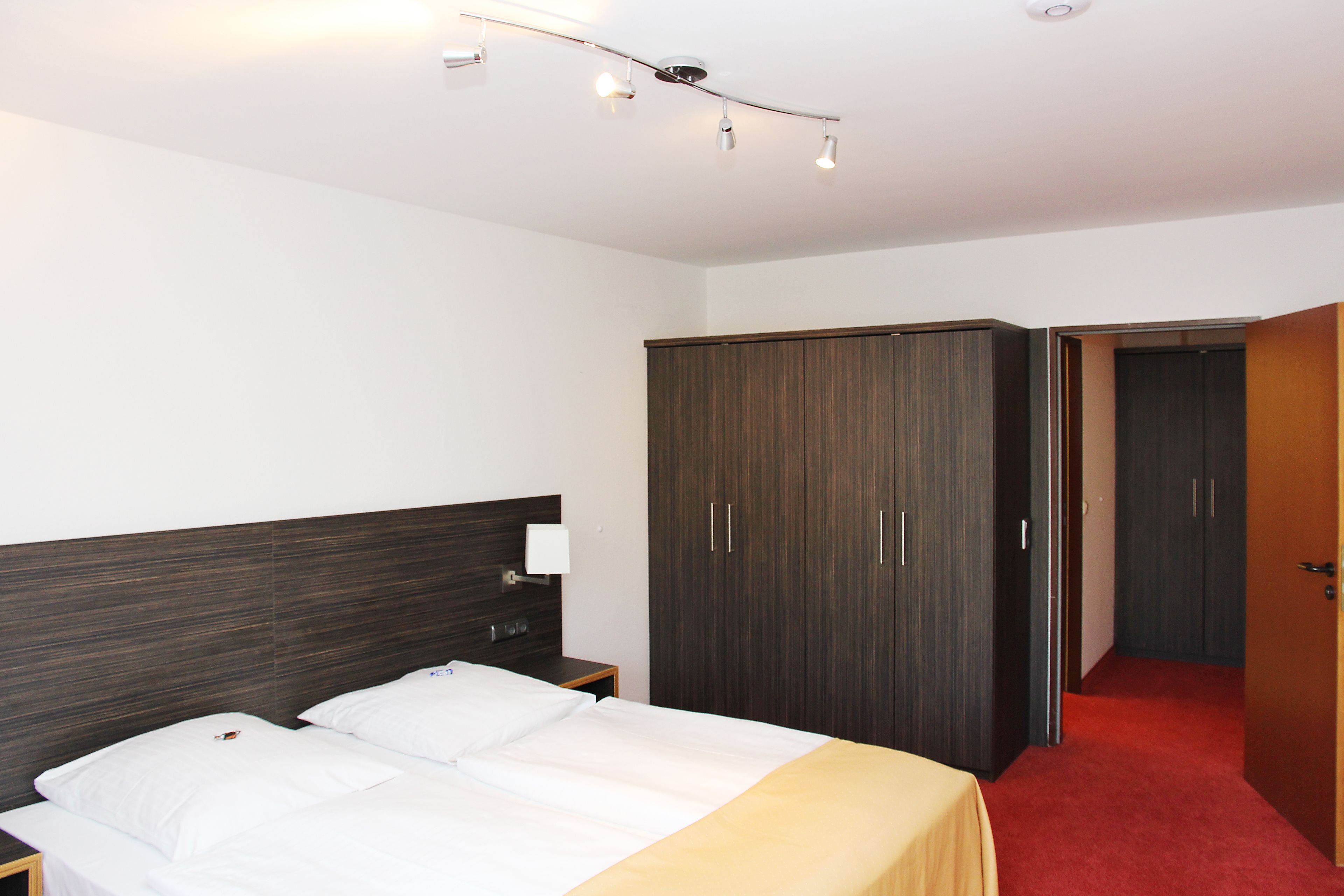 Hotellihuone, jossa on sänky, vaatekaappi ja punaisella matolla päällystetty käytävä.