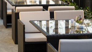 Outdoor dining tables with wicker chairs and cushioned seats, arranged along a waterfront promenade.