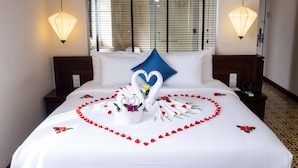 A neatly made bed with a heart-shaped arrangement of rose petals and swan decorations, a blue pillow, and two bedside lamps.