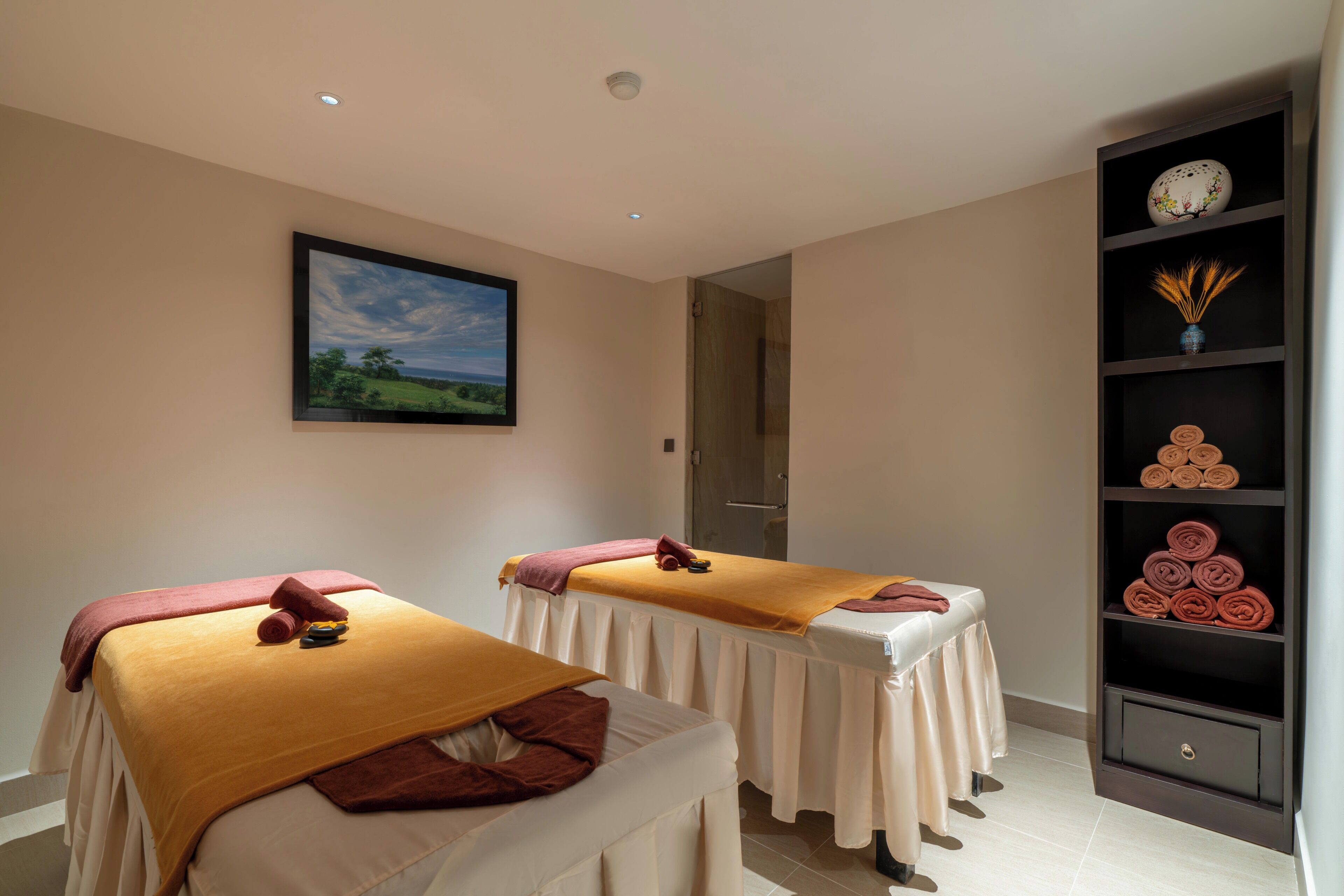 A spa room with two massage tables, a painting of a landscape, and a shelf with rolled towels.