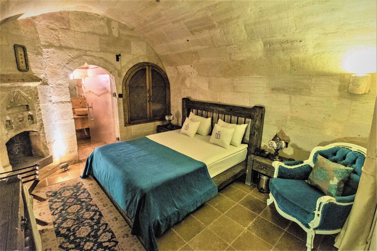 A bedroom with a stone wall, a bed with a blue bedspread, a wooden nightstand, a chair, and a small table with a lamp.