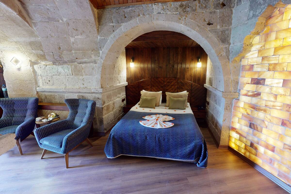A room with a stone wall, a wooden ceiling, a bed with a patterned bedspread, two patterned chairs, and a wall with a mosaic design.