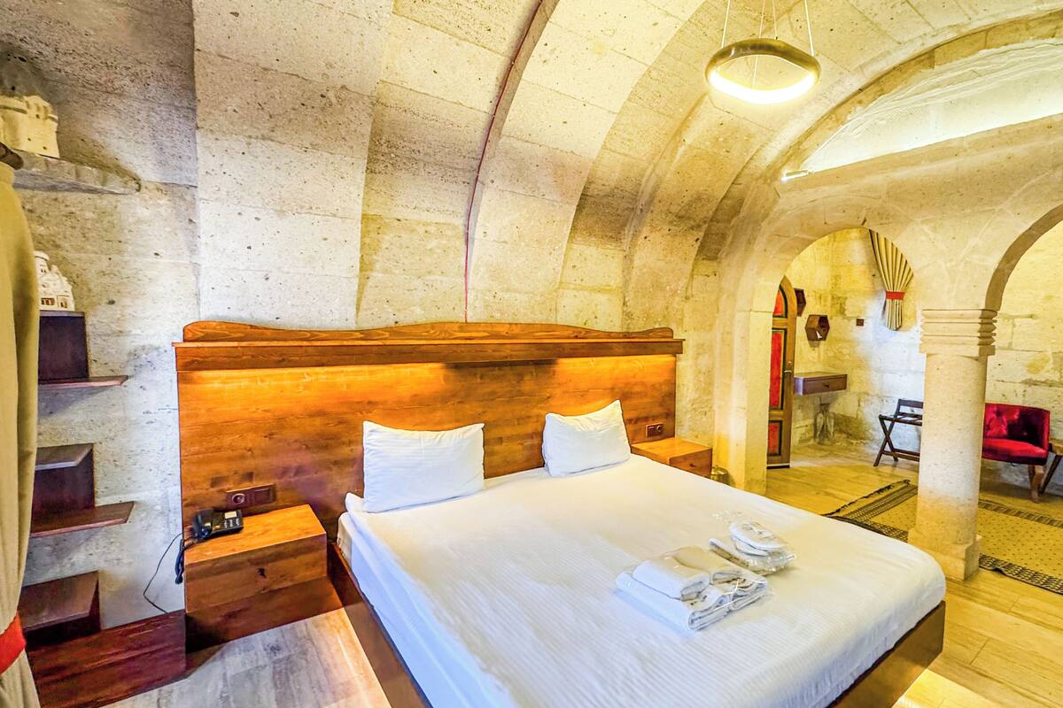 A room with a large bed, wooden headboard, and a stone wall. There is a small table with a lamp and a chair.