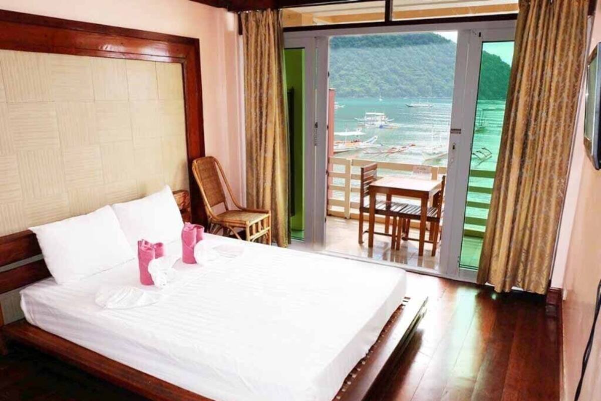 Ein ordentlich bezogenes Bett mit weißen Leinen und rosa Handtüchern, ein Holz-Kopfteil und ein Balkon mit Blick auf Boote und Grünflächen.