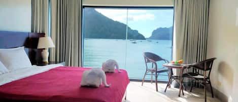 A hotel room with a large bed, a small table with chairs, and a view of the sea through a large window.
