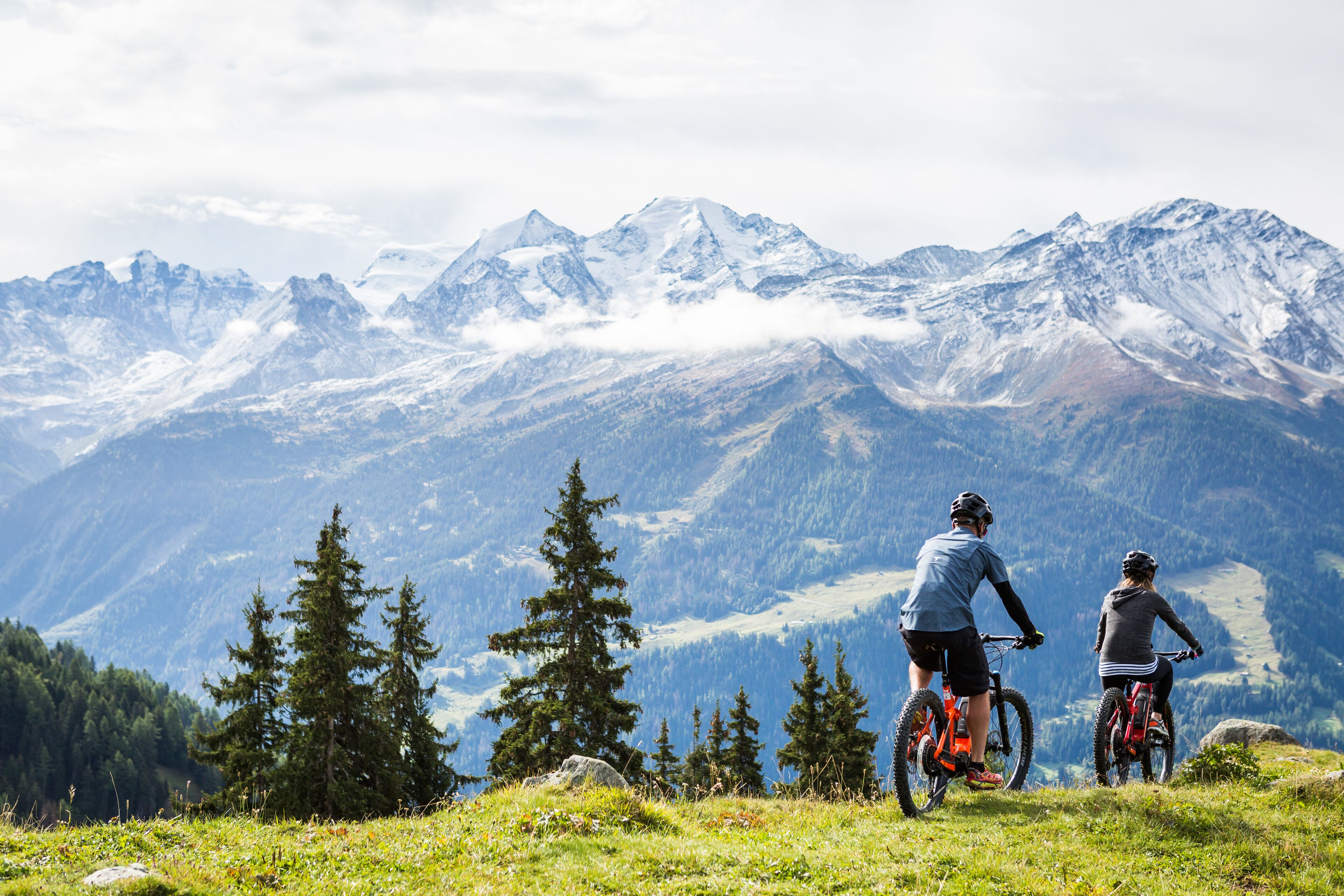 Dos personas en bicicletas de montaña en una colina cubierta de hierba, con vistas a montañas nevadas.