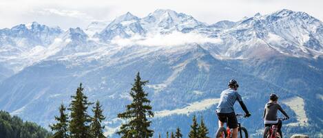 Dos personas en bicicletas de montaña en una colina cubierta de hierba, con vistas a montañas nevadas.