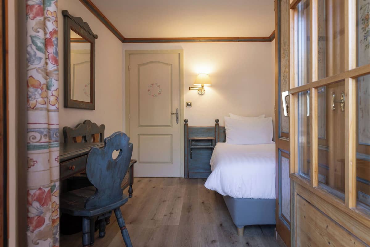 Un dormitorio con un escritorio de madera, una silla, una cama con ropa blanca y una puerta adornada con un adorno, además de una lámpara fijada a la pared.