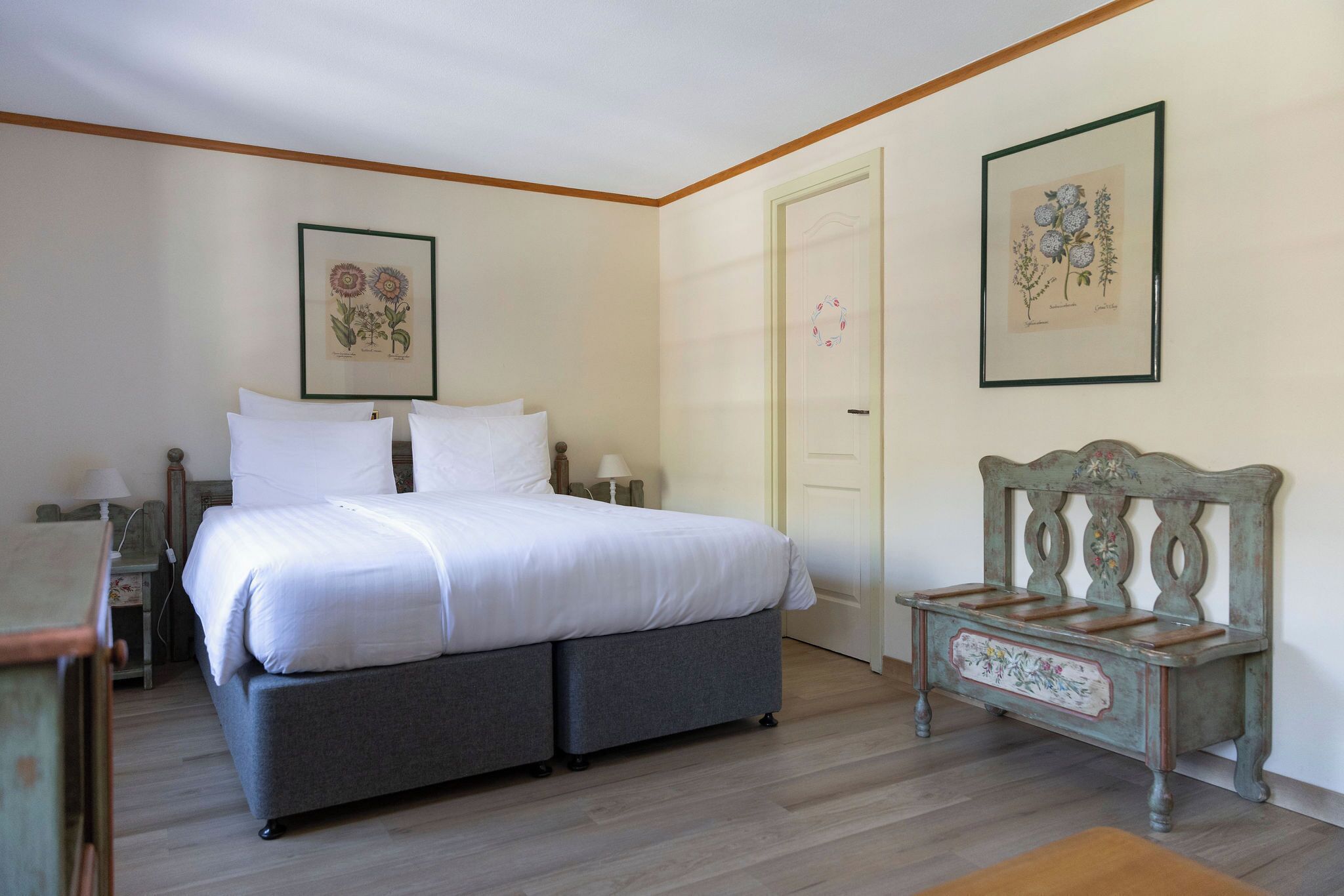 A bedroom with a double bed, a bench, and two framed botanical illustrations on the walls.