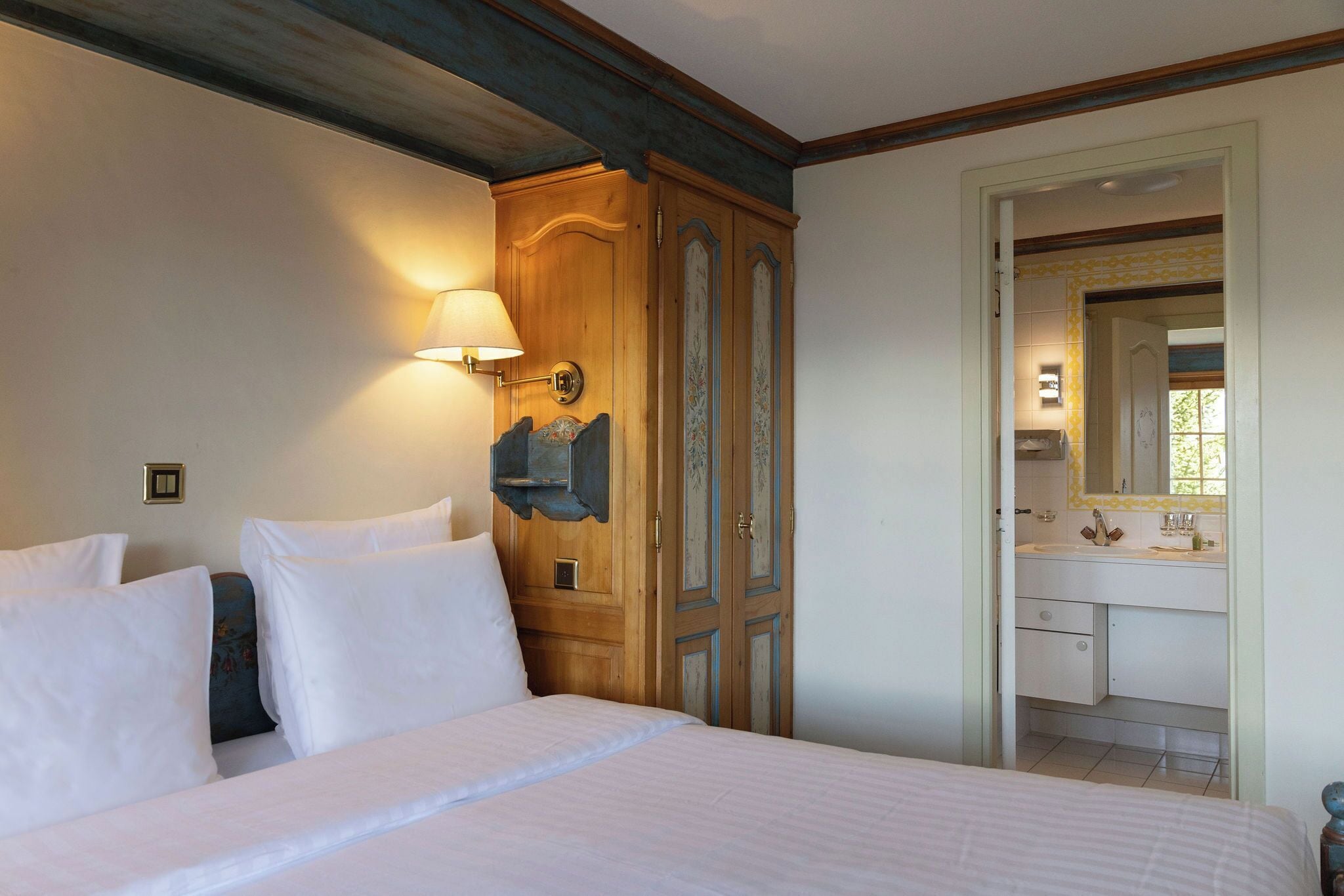 Quarto de hotel com cama, armário de madeira, banheiro com espelho e luminária na parede.