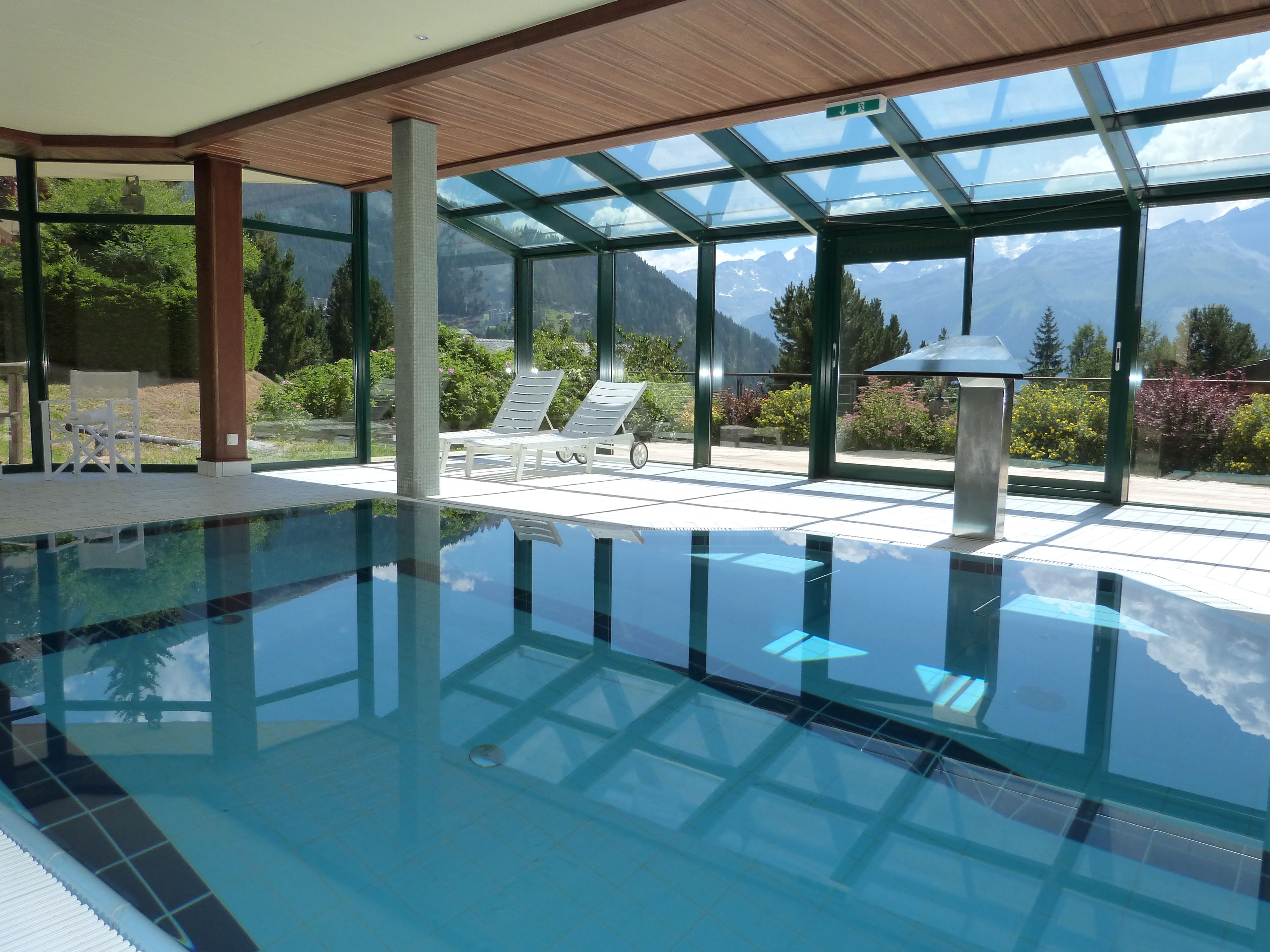 An indoor swimming pool with clear glass walls, lounge chairs, and a view of mountains.