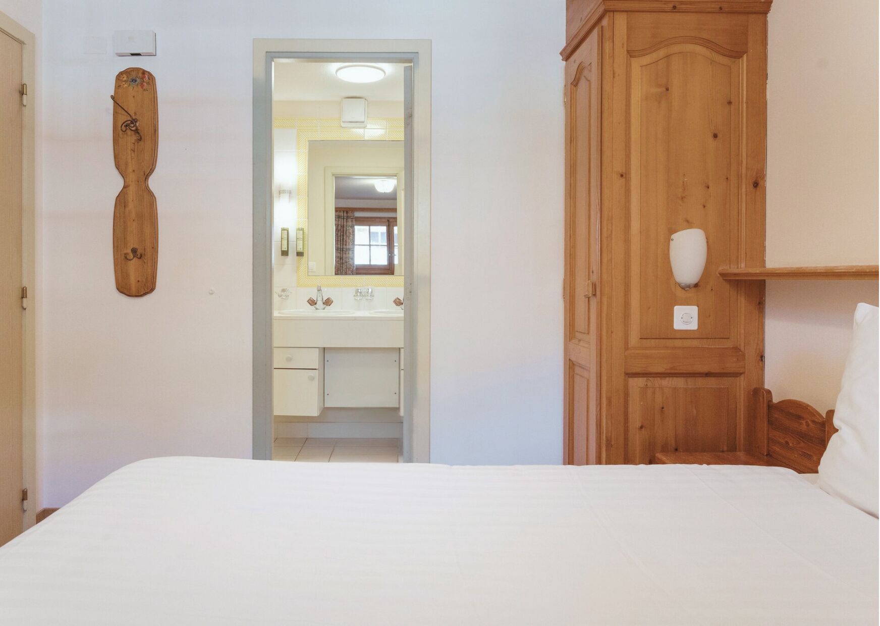 A bedroom with a wooden wardrobe, a bed, and a bathroom visible through an open door.