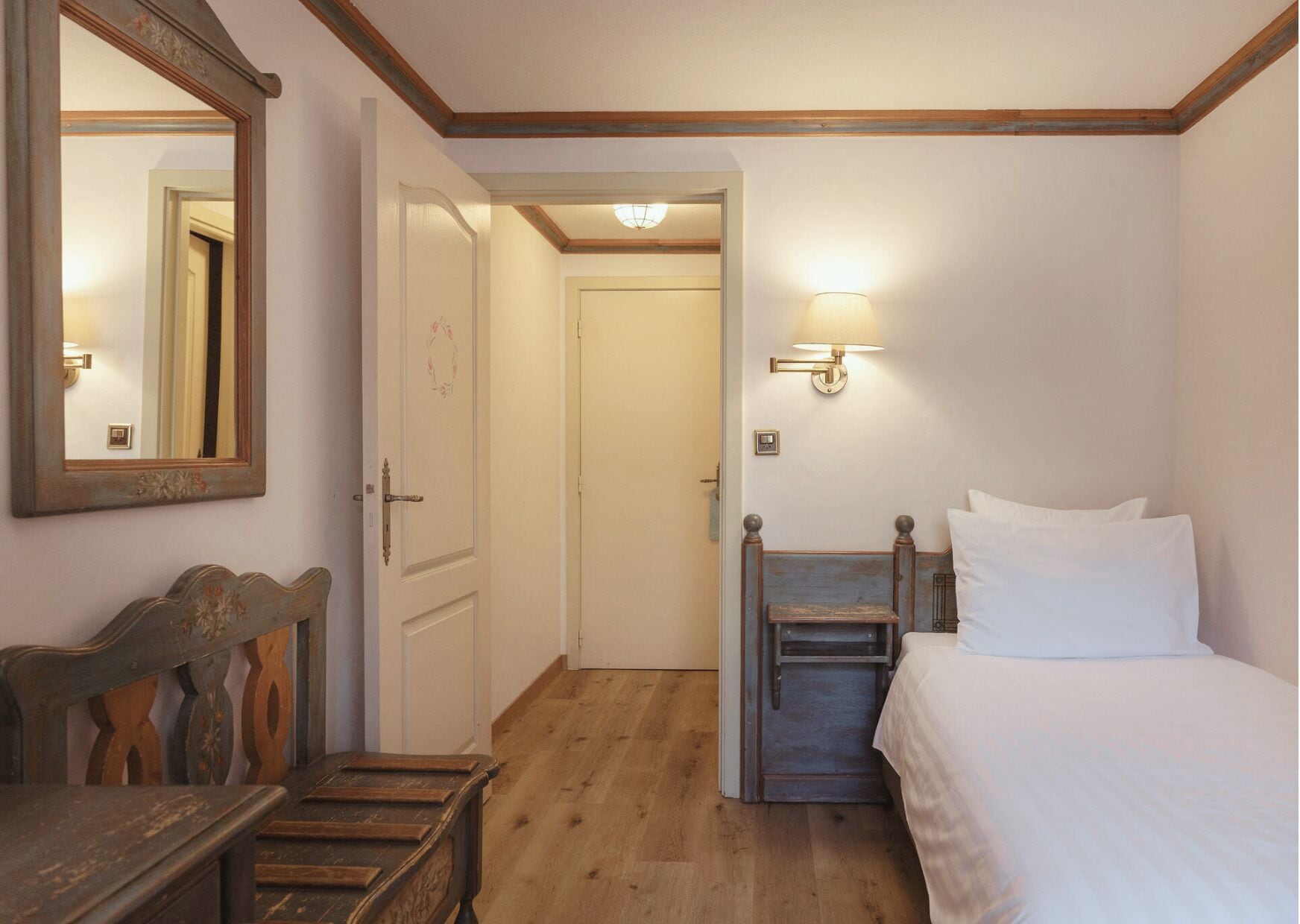 A bedroom with a wooden bench, a bed with white linens, a wall-mounted lamp, and a door leading to another room.