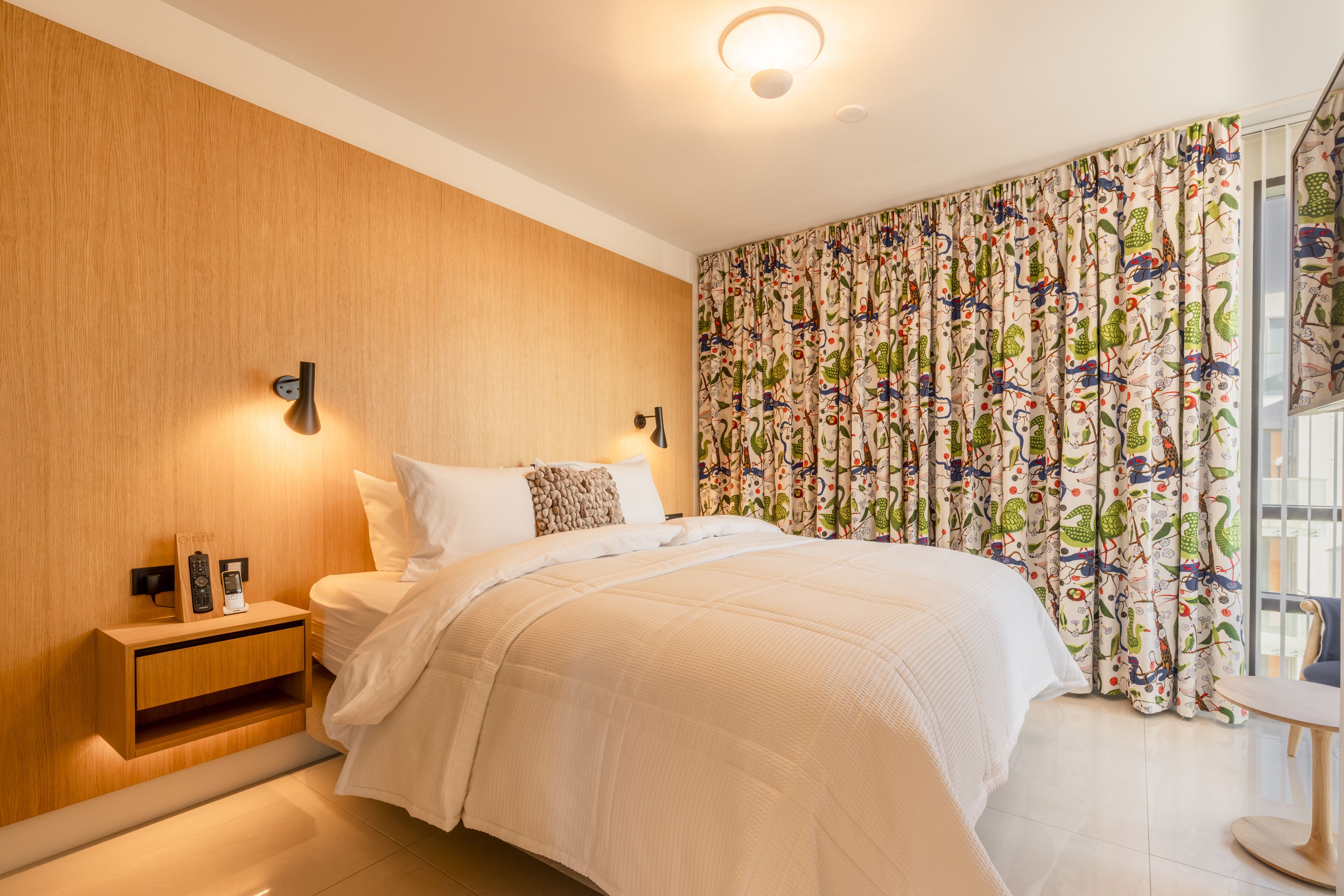 A bedroom with a wooden headboard, a bedside table with a phone and a remote, a bed with white bedding, and a colorful curtain.