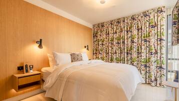 A bedroom with a wooden headboard, a bedside table with a phone and a remote, a bed with white bedding, and a colorful curtain.