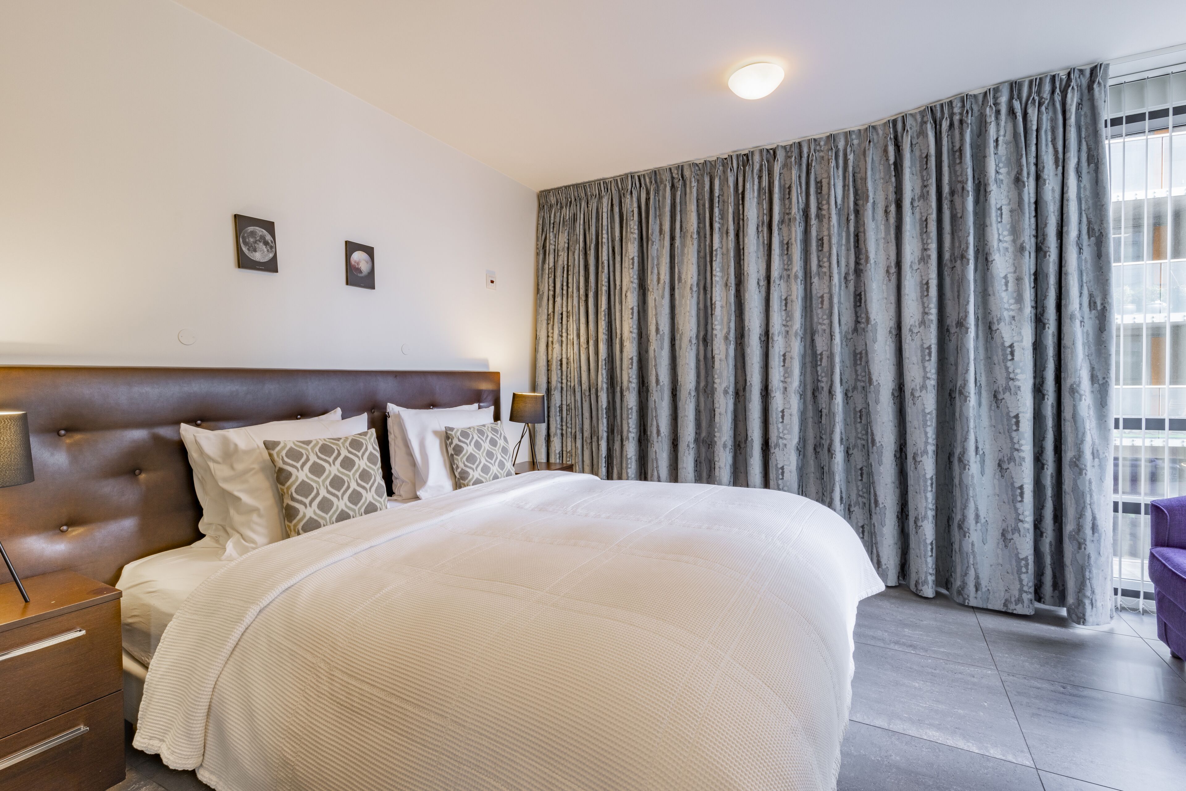 Habitación de hotel con una cama grande, cabecera de madera, mesita de noche con lámpara y vistas al exterior a través de cortinas.