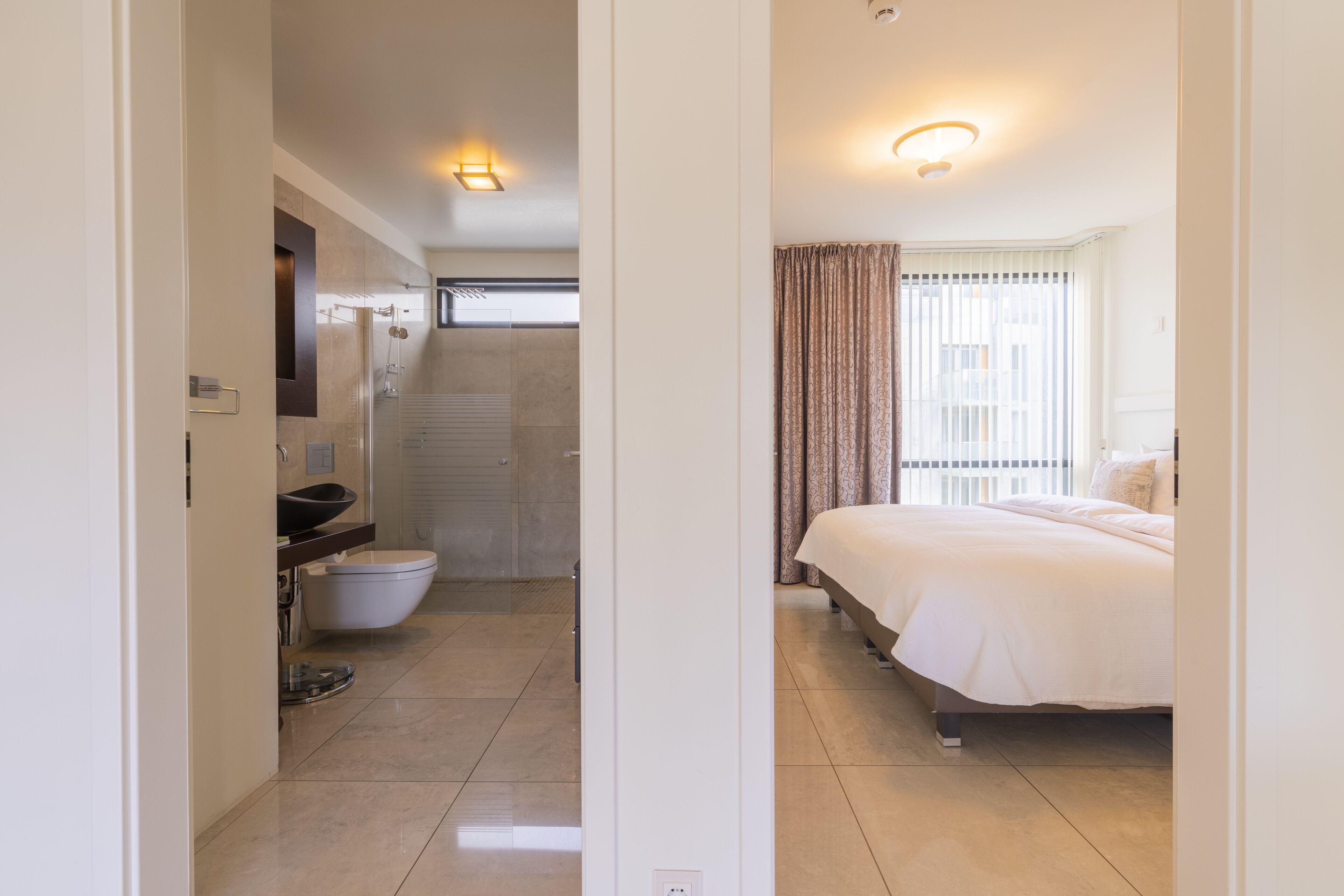 A modern hotel room with a bathroom, a shower, a bed, and a window with curtains.