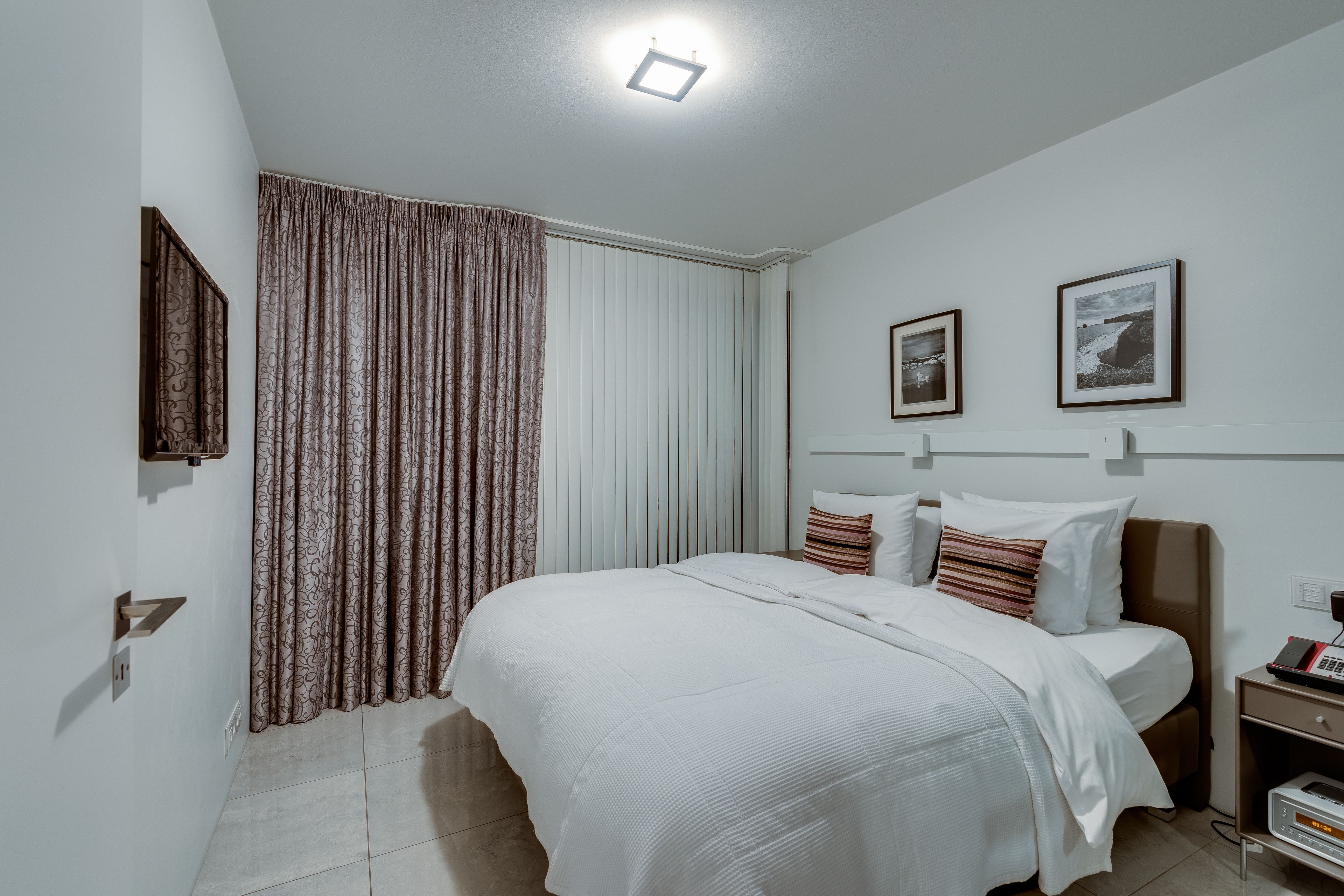 Un dormitorio moderno con una cama grande, una mesita de noche, un televisor montado en la pared y un ventanal con cortinas.