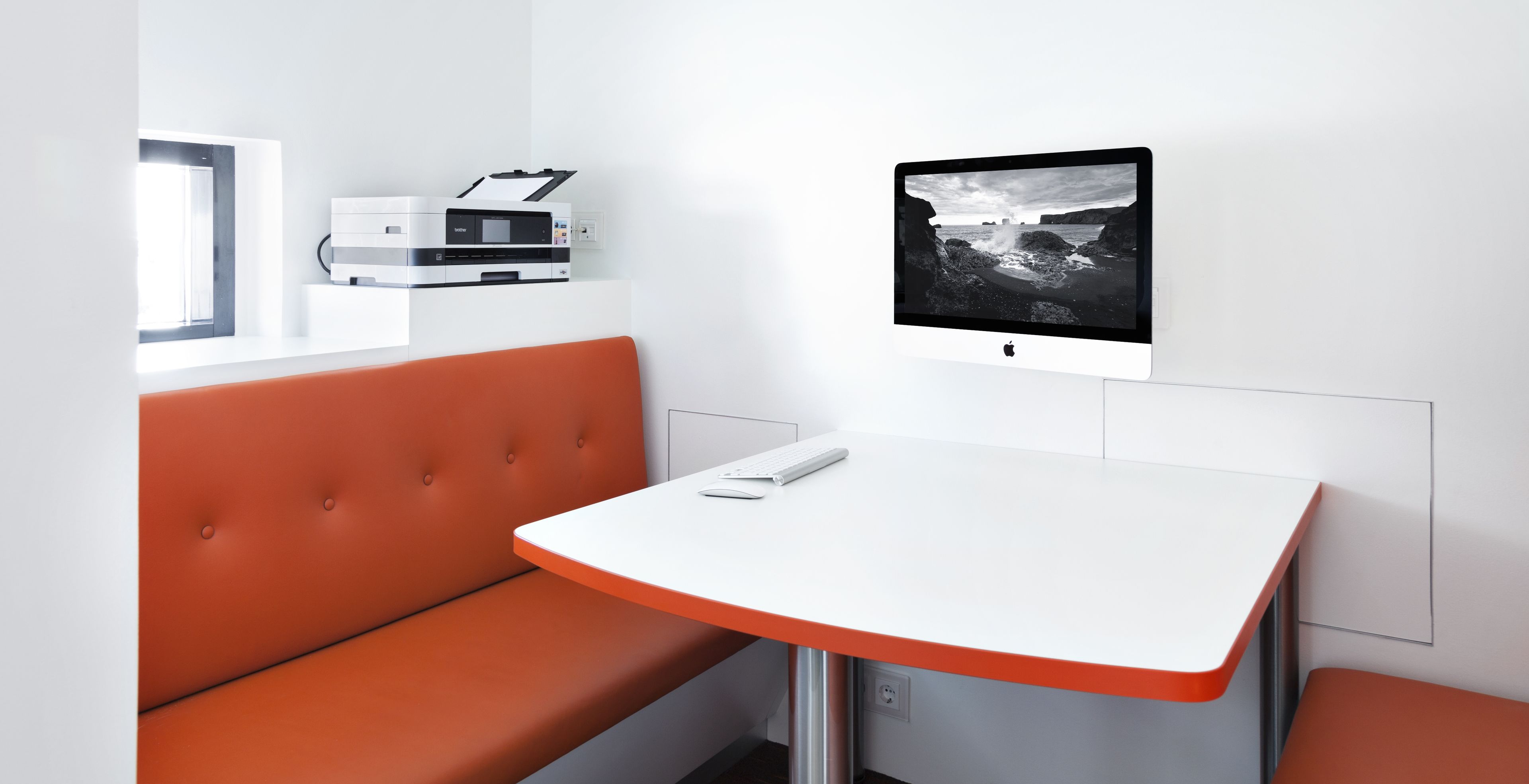 A modern room with a white table, orange seating, a printer, and a monitor displaying a landscape.