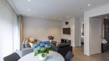 Una sala moderna con un sofá, una mesa de centro y una mesa de comedor con un jarrón de flores. Hay un televisor de pantalla plana montado en la pared y una puerta que conduce a un baño.