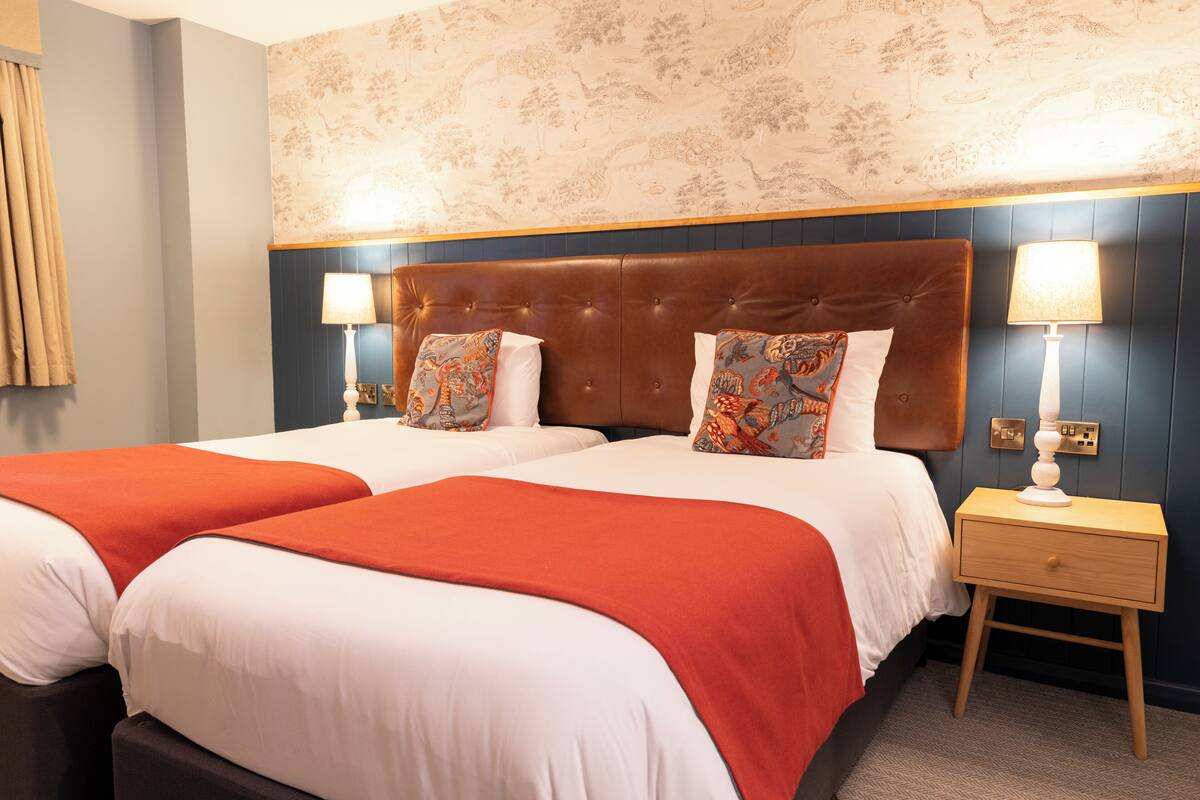 A hotel room with two beds, a nightstand, and a wall with floral wallpaper.