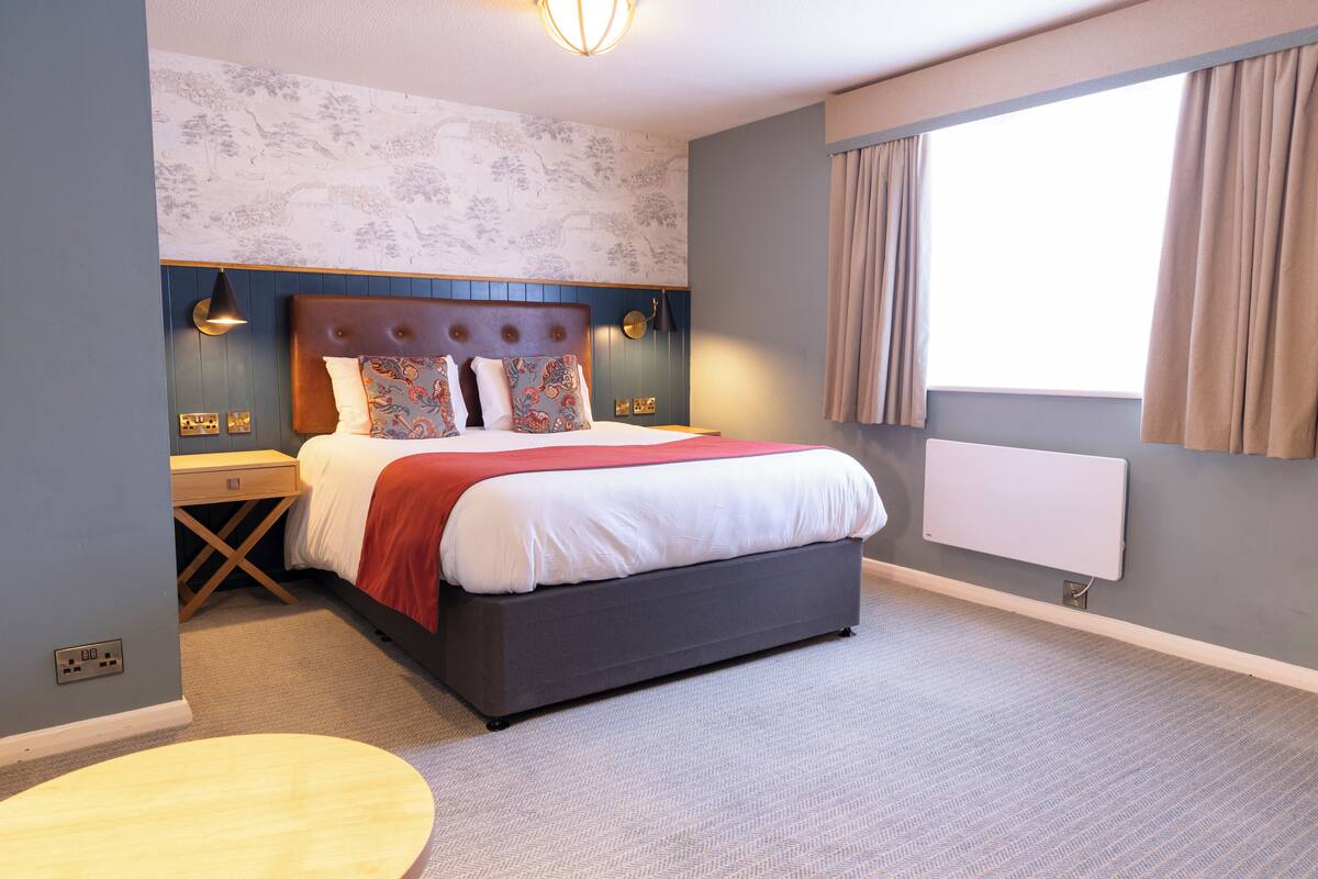 A modern hotel room with a large bed, a bedside table, a wall-mounted heater, and a window with curtains.