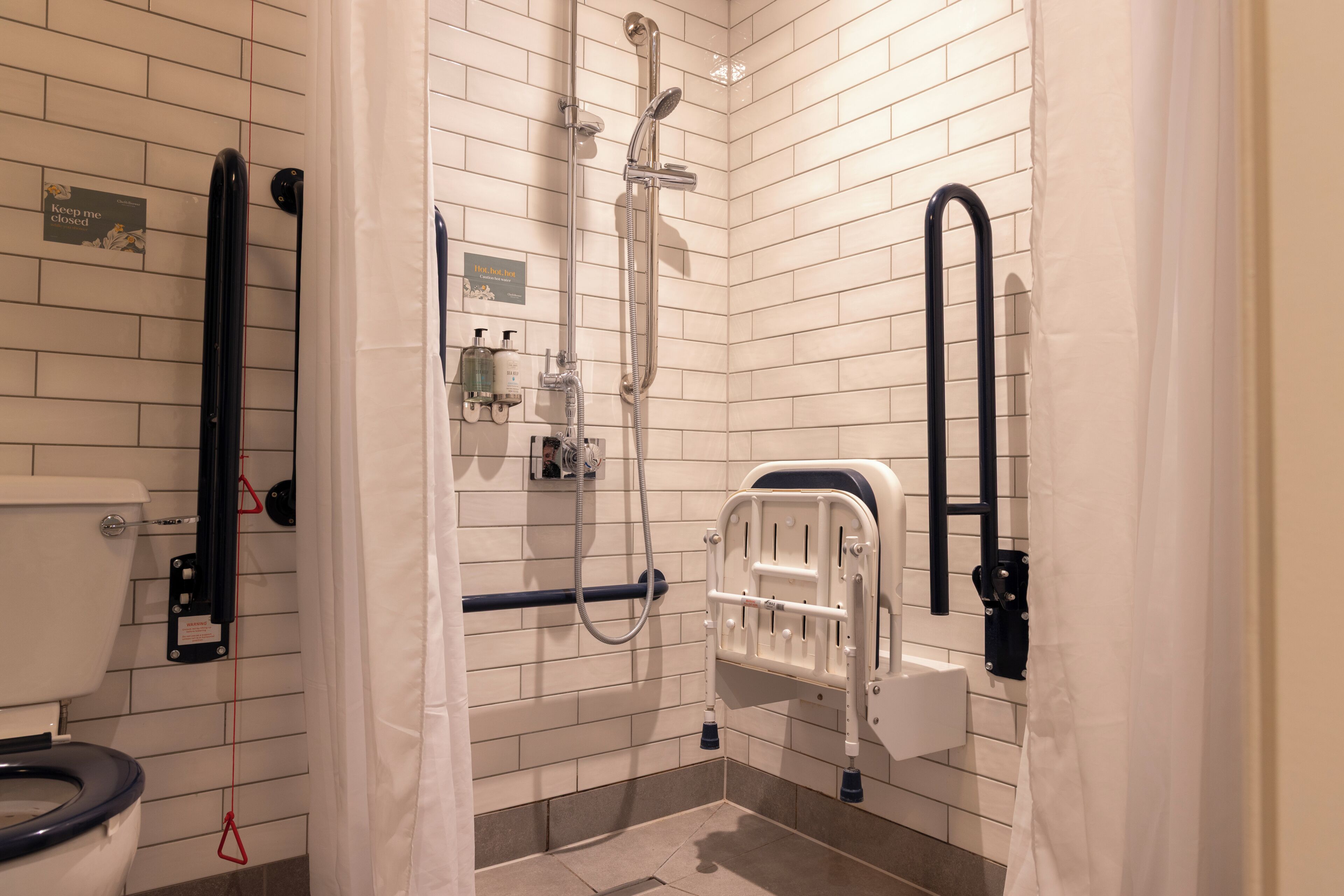 A bathroom with white tiled walls, a shower with a handheld showerhead, a toilet with a black seat, and a grab bar near the toilet.