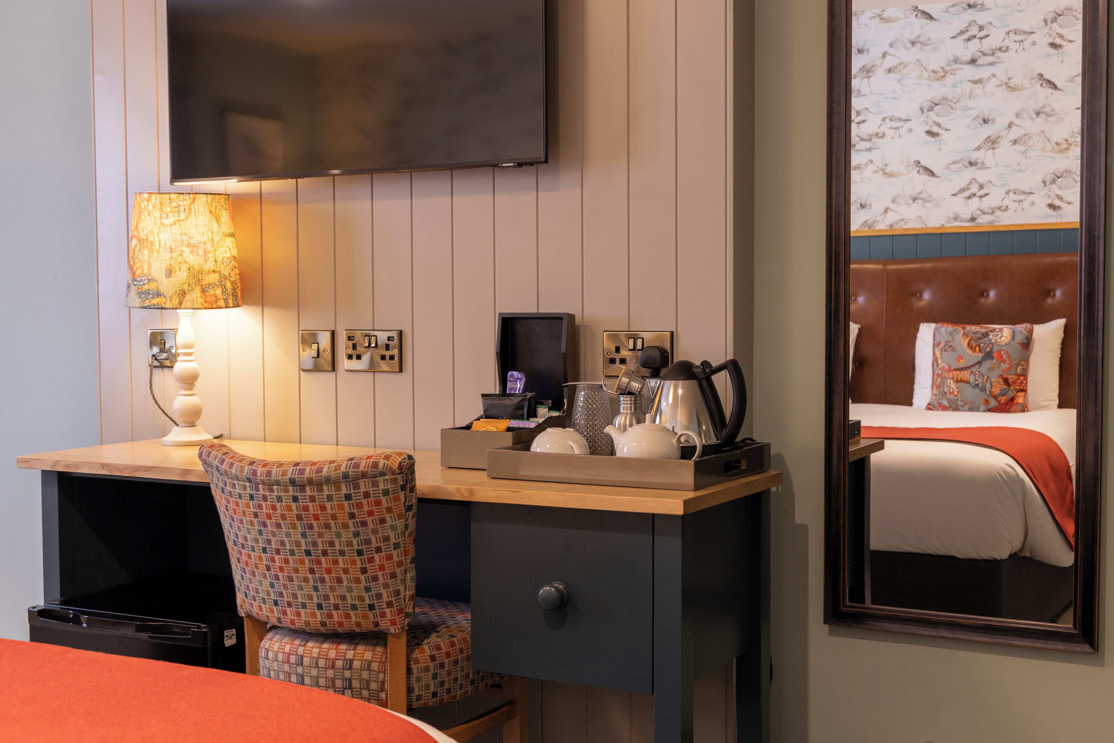Habitación de hotel con televisión de pantalla plana, escritorio con lámpara, silla, cafetera y cama con cabecera estampada.