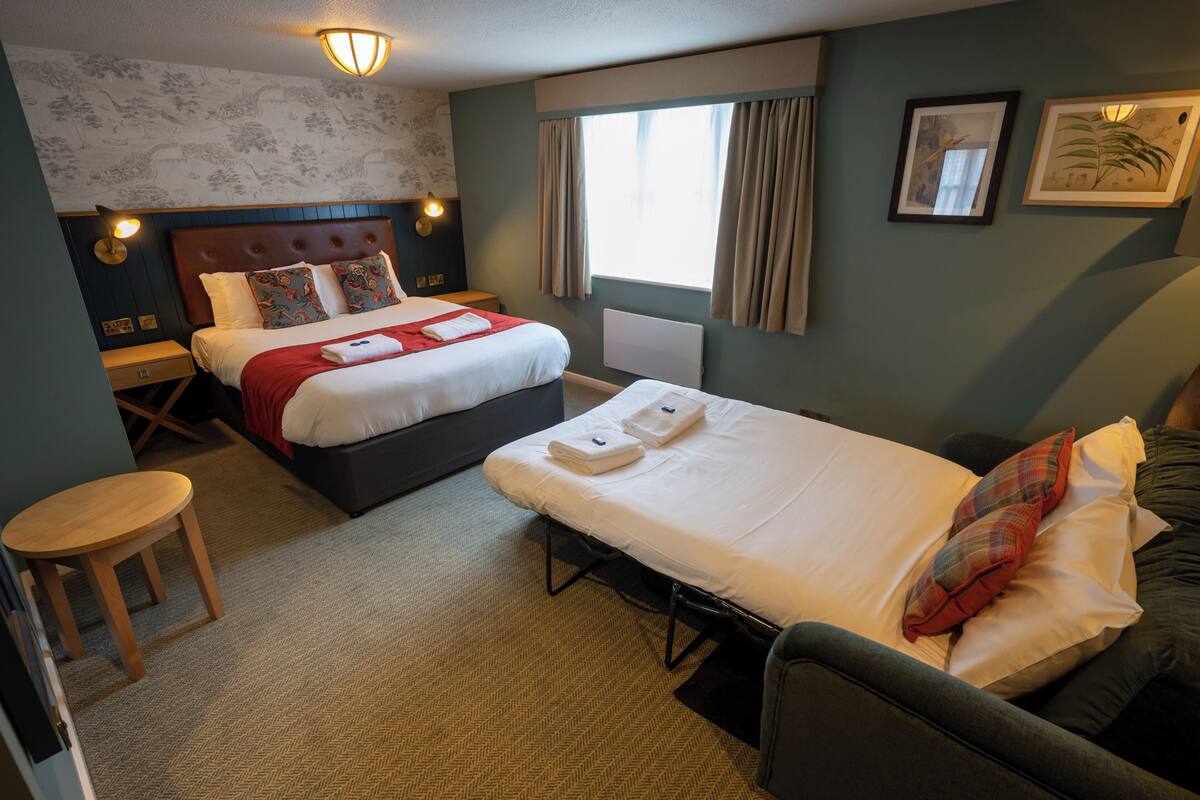 A hotel room with two beds, a small table, a nightstand, and a window with curtains.