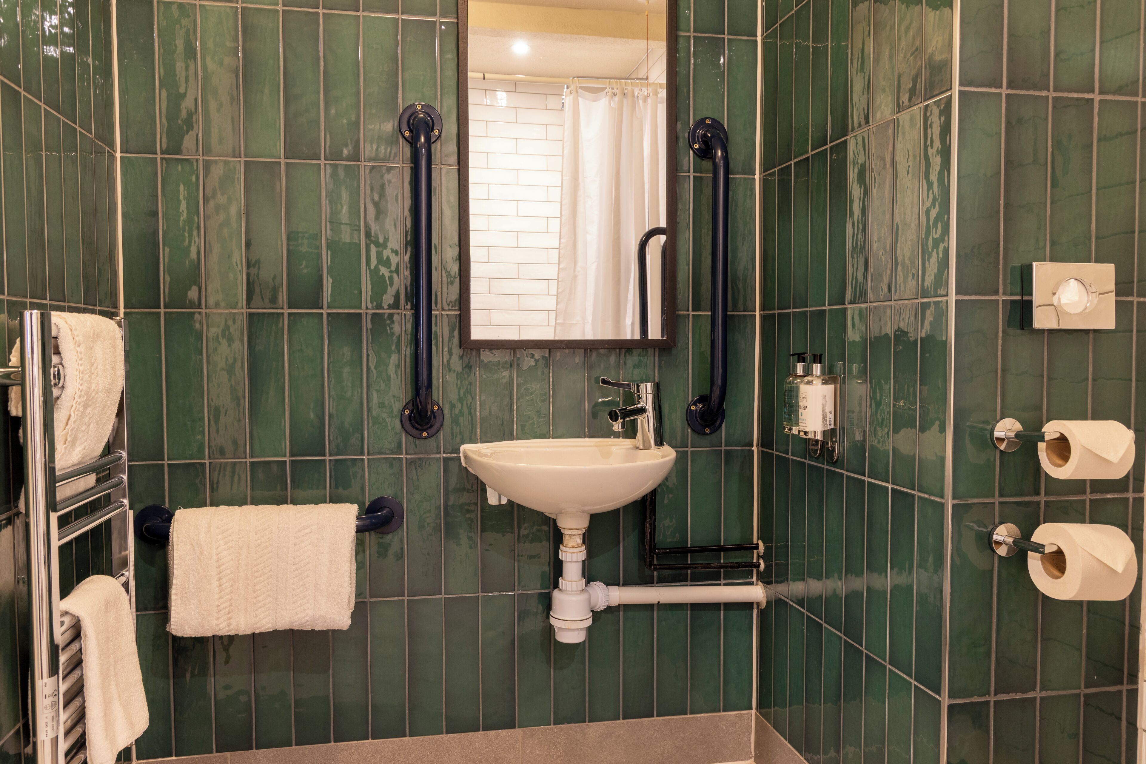 Un baño con lavamanos, espejo, toallero y dispensador de papel higiénico.