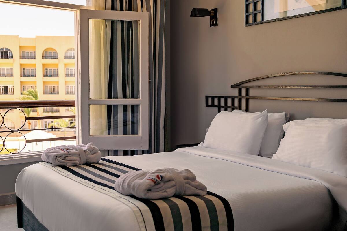 Habitación de hotel con cama, ventana con cortinas, vista de un edificio y una lámpara fijada a la pared.