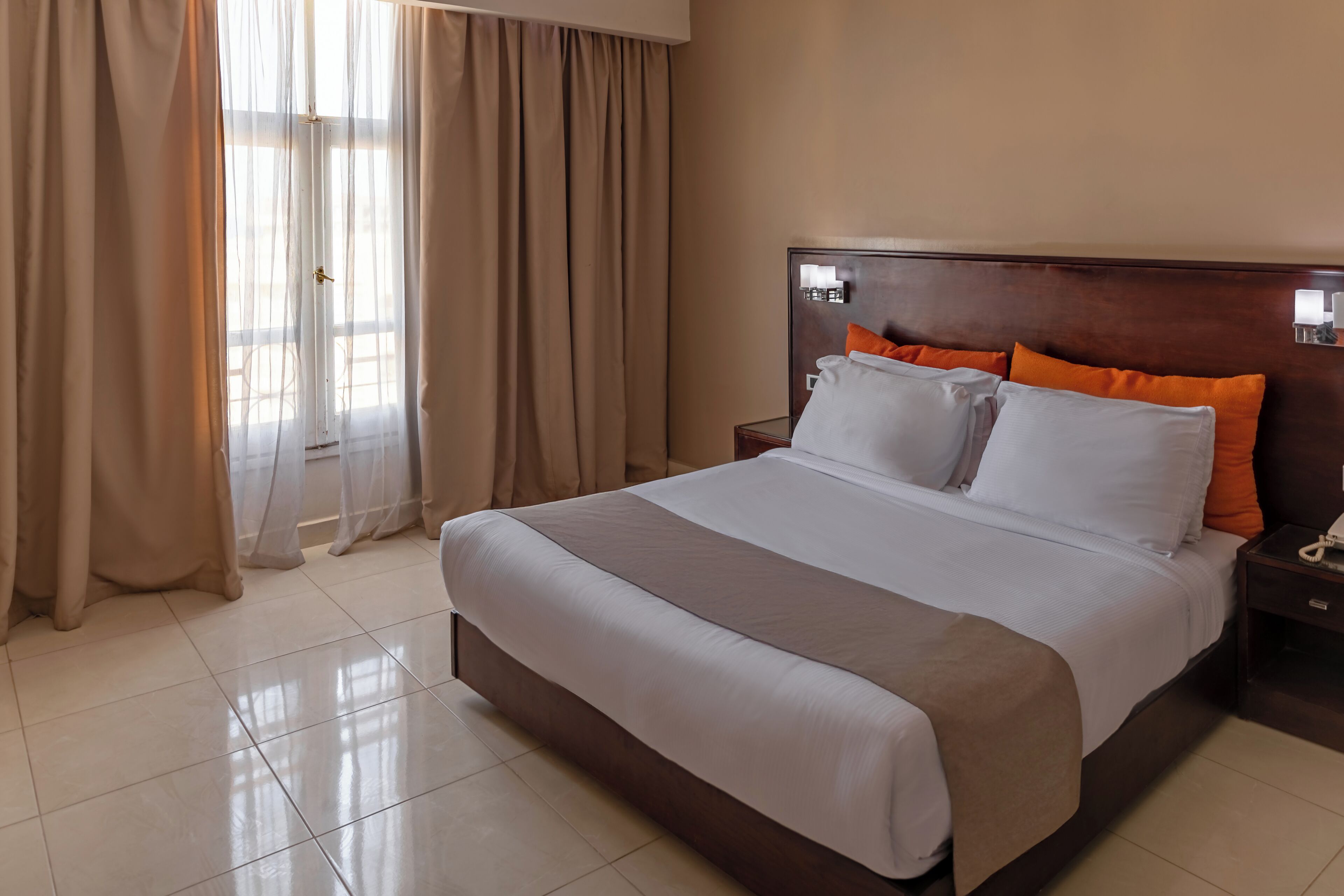 Una habitación de hotel con una cama grande, una mesita de noche con teléfono y cortinas translúcidas en la ventana.