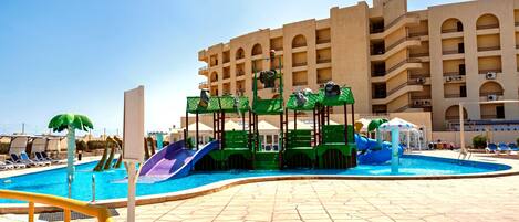 Una piscina con toboganes y zona de juegos acuáticos, rodeada por un edificio con varios balcones.