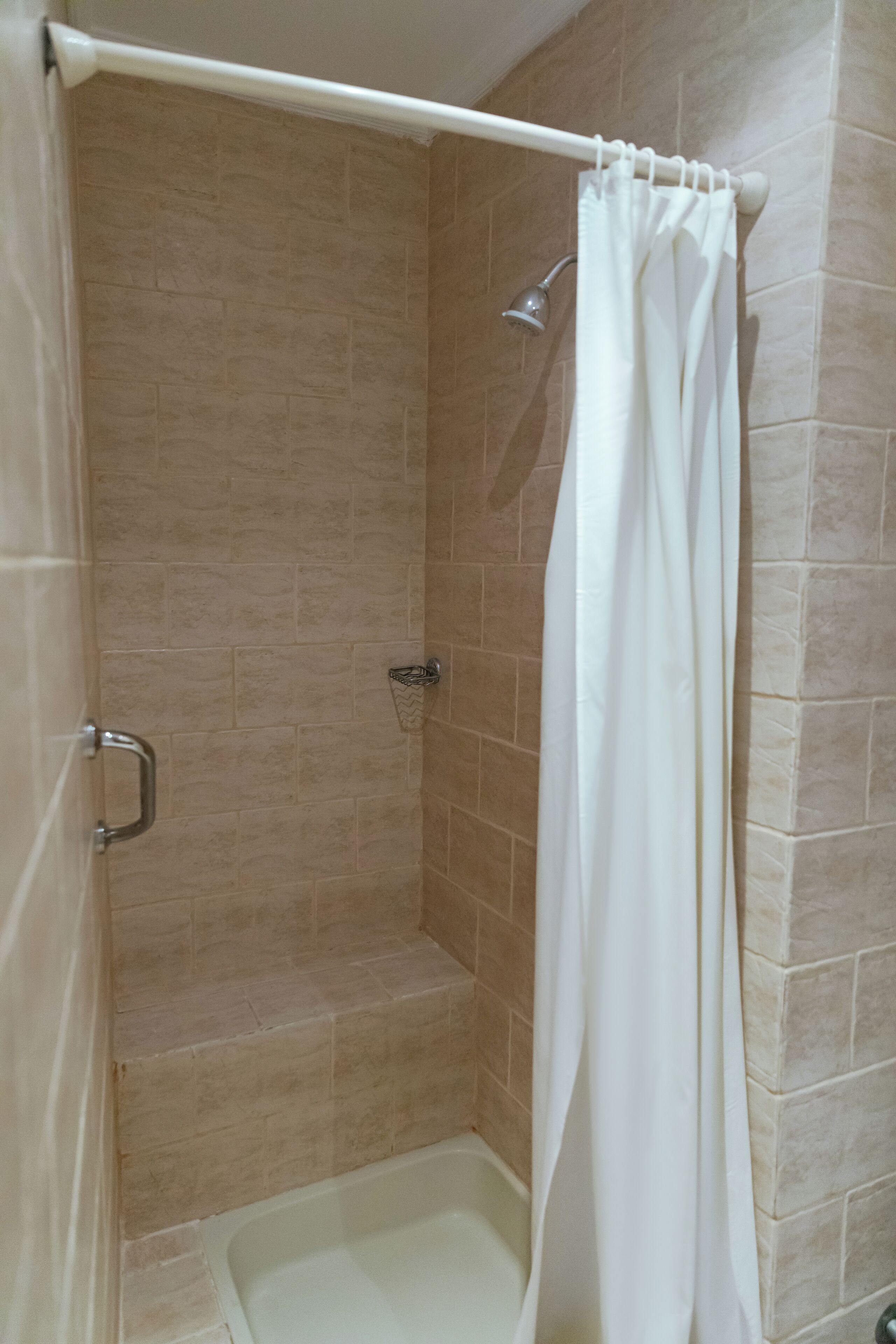 Ducha con cortina blanca, paredes de azulejos color beige y un cabezal de ducha de mano.