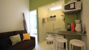 Een knus woongedeelte met een kitchenette, een bruine bank met kussens, en een klein barretje met krukjes.