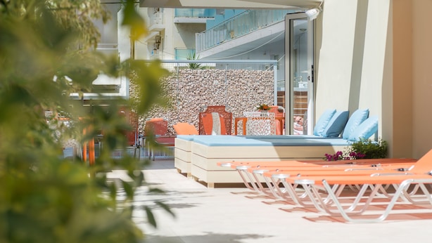 A sunlit outdoor seating area with a sofa and chairs, a stone wall, and a view of a building with balconies. - Majestic (Rhodes)