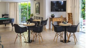 A modern indoor space with a pool table, multiple seating areas, a flat-screen TV, and large windows offering a view of greenery. - Majestic (Rhodes)